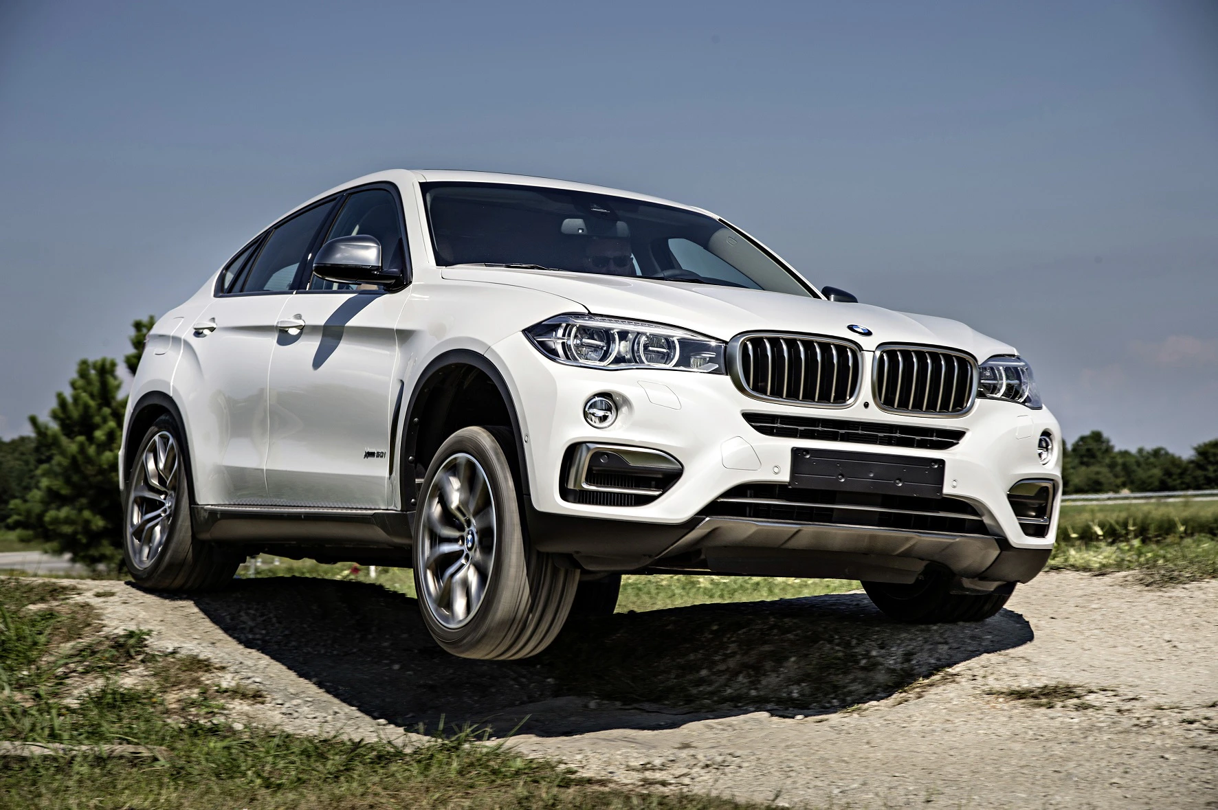Ein weißer BMW-SUV fährt über einen unbefestigten Weg, vor blauem Himmel und grüner Landschaft.