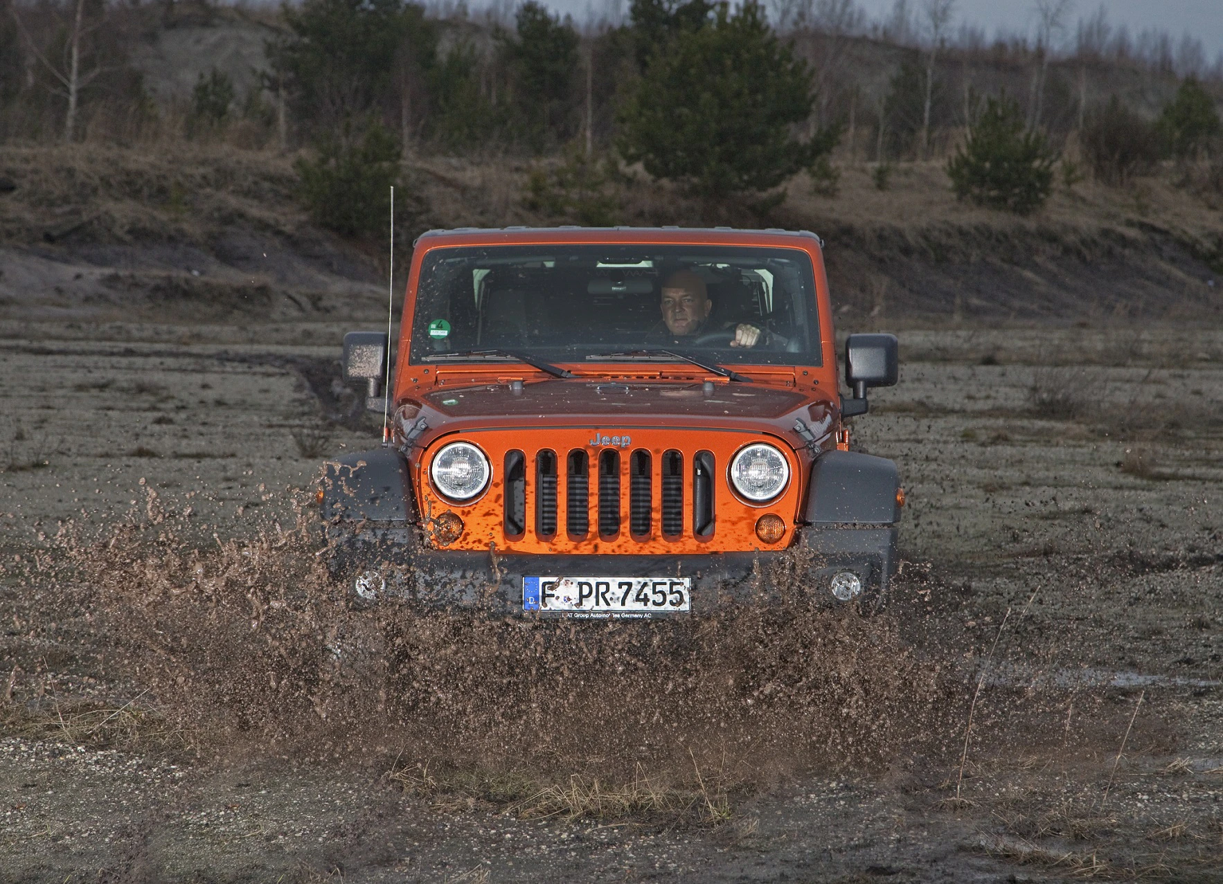 Oranger Jeep fährt durch schlammiges Gelände, aufgewühlte Erde spritzt seitlich hoch. Im Hintergrund sind Bäume und ein bewölkter Himmel zu sehen.