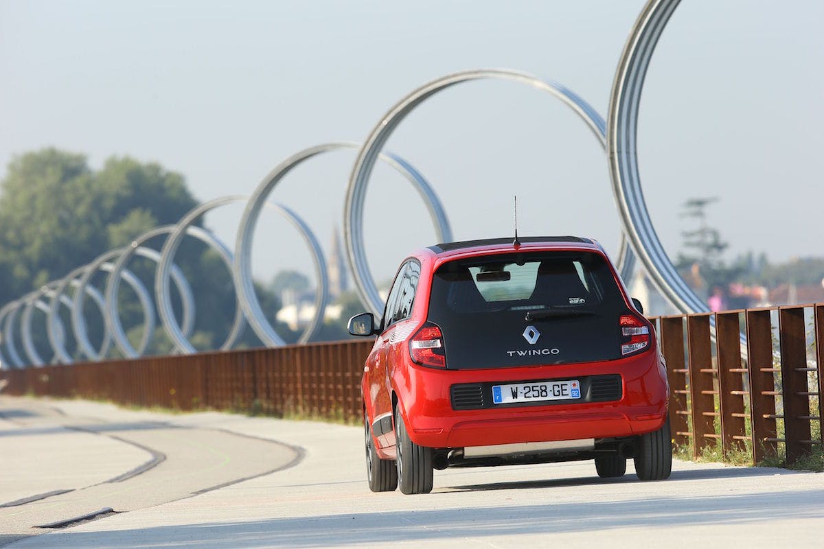 Roter Renault Twingo fährt auf einer Straße mit kunstvollen Metallbögen im Hintergrund. Klarer Himmel und vereinzelte Bäume am Straßenrand.