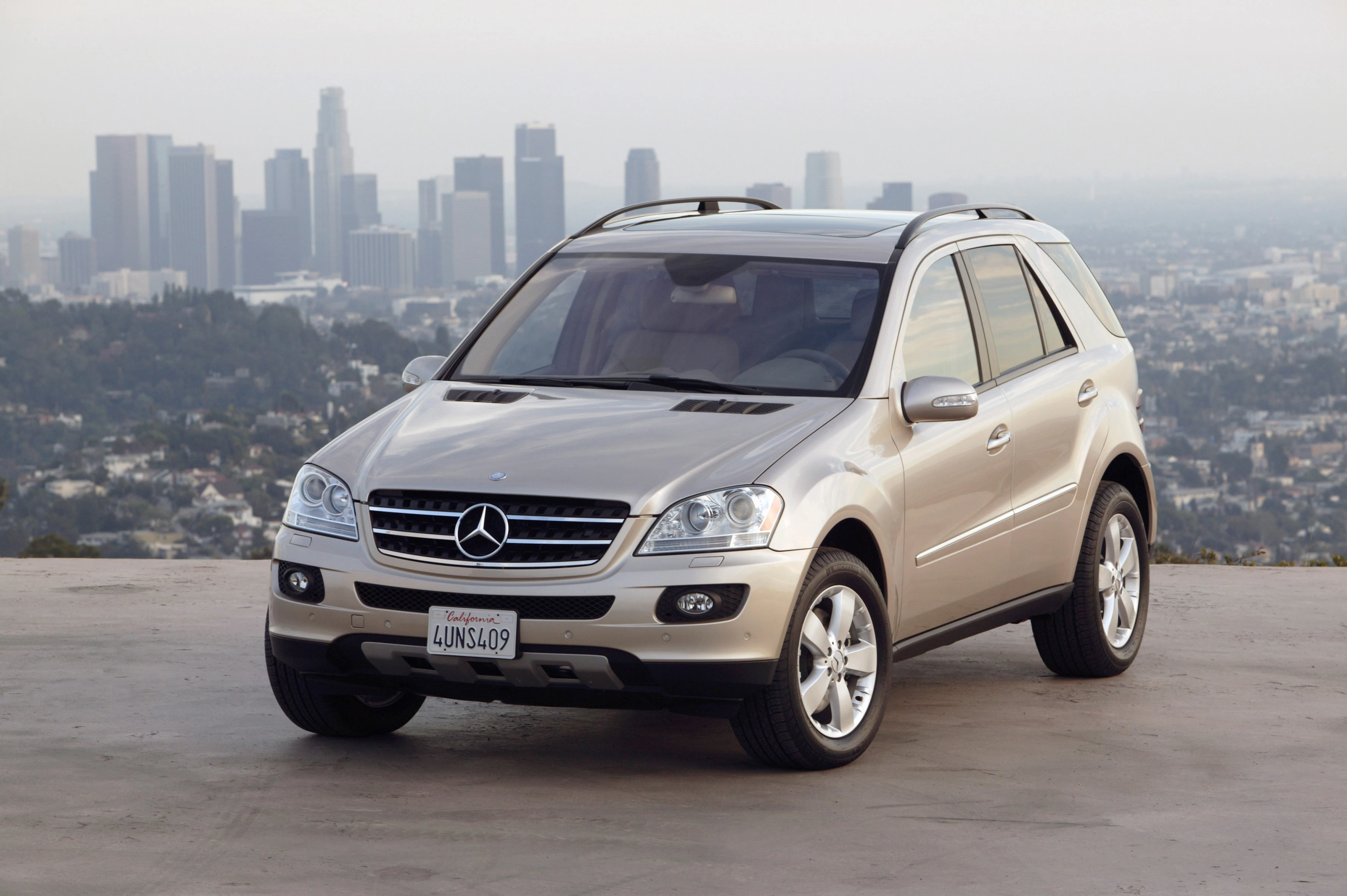 Ein silberner Mercedes SUV parkt auf einer Anhöhe mit einer verschwommenen Stadtansicht im Hintergrund.
