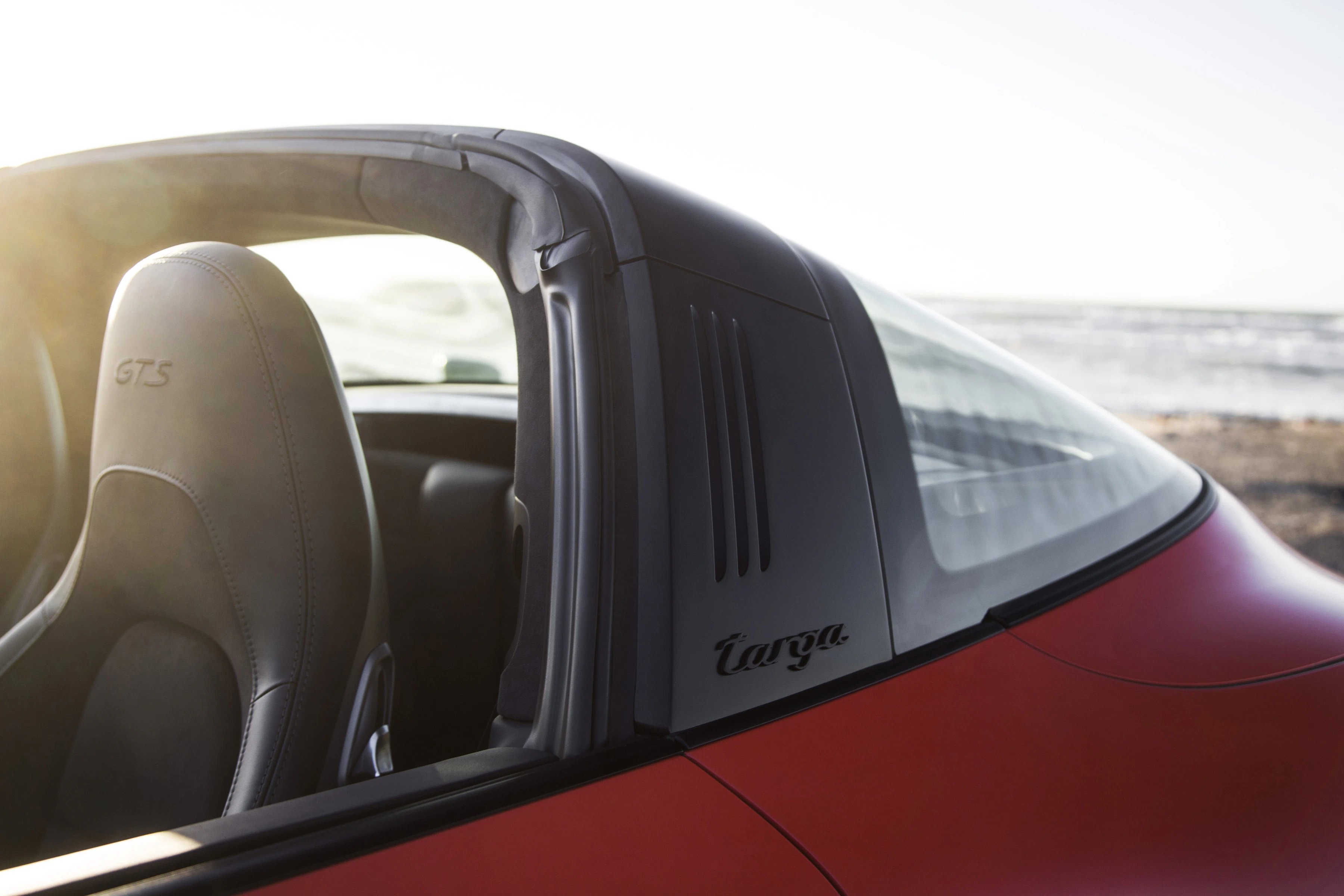 Roter Sportwagen mit Targa-Überrollbügel, offenem Verdeck und schwarzem Ledersitz vor einer Küstenlandschaft bei Sonnenuntergang.