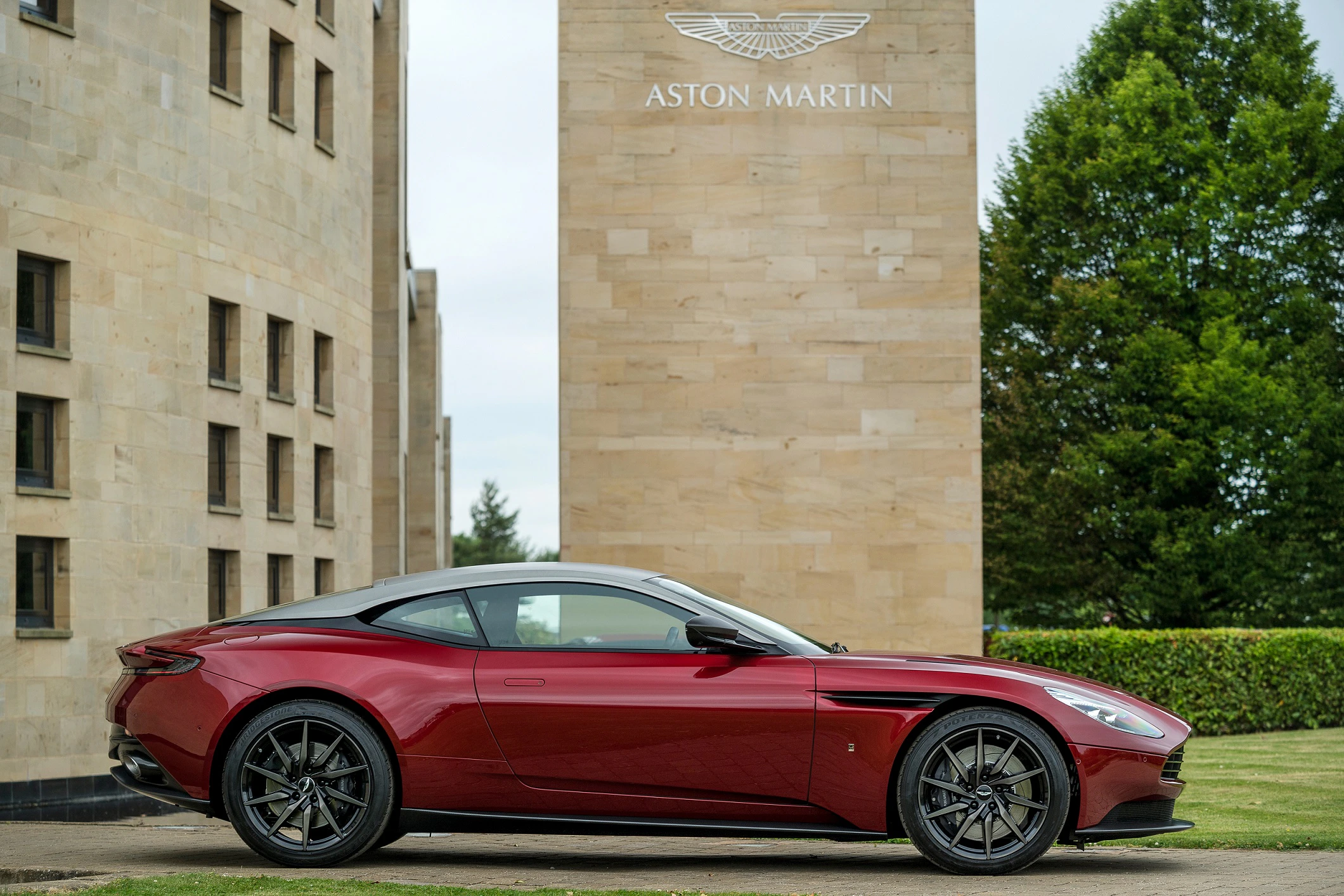 Roter Aston Martin Sportwagen vor einem modernen, beige-grauen Gebäude mit Bäumen im Hintergrund.