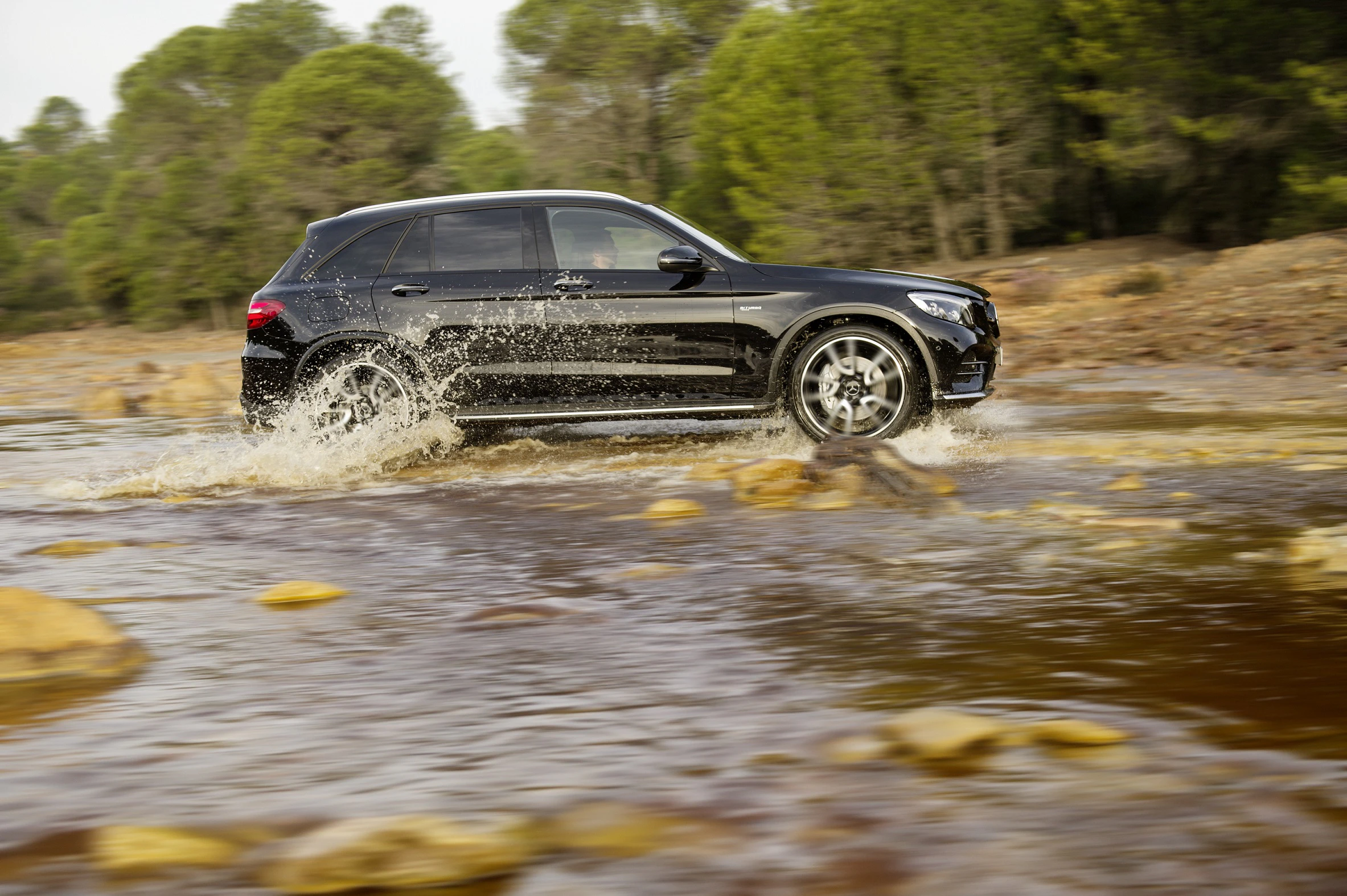 Schwarzer SUV fährt durch flaches Wasser in einer bewaldeten Landschaft, spritzt Wasser umher, während es Gelände überquert.