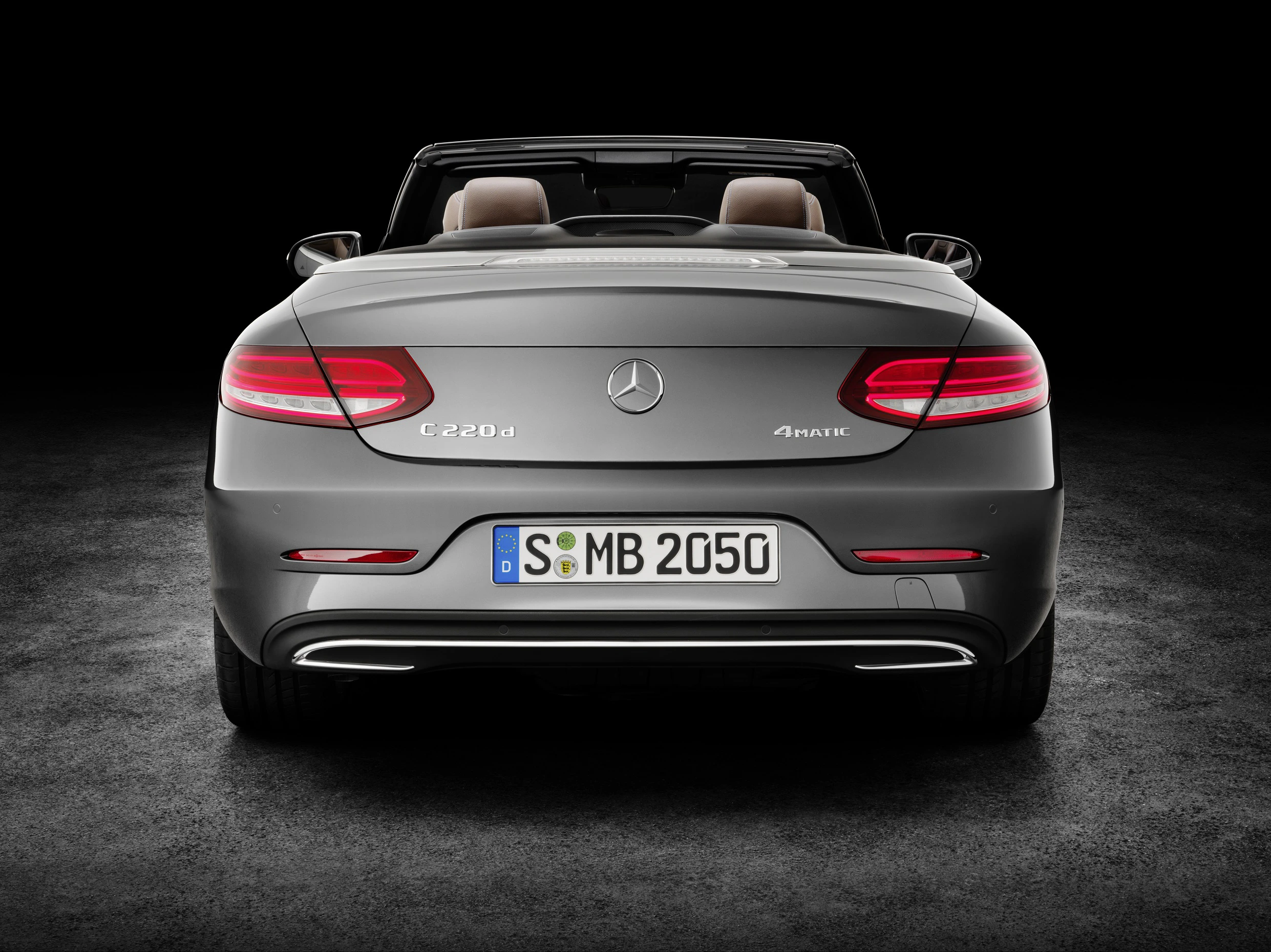 Rückansicht eines grauen Mercedes C220d 4MATIC Cabriolets auf schwarzem Hintergrund. Heckleuchten leuchten, Nummernschild sichtbar.