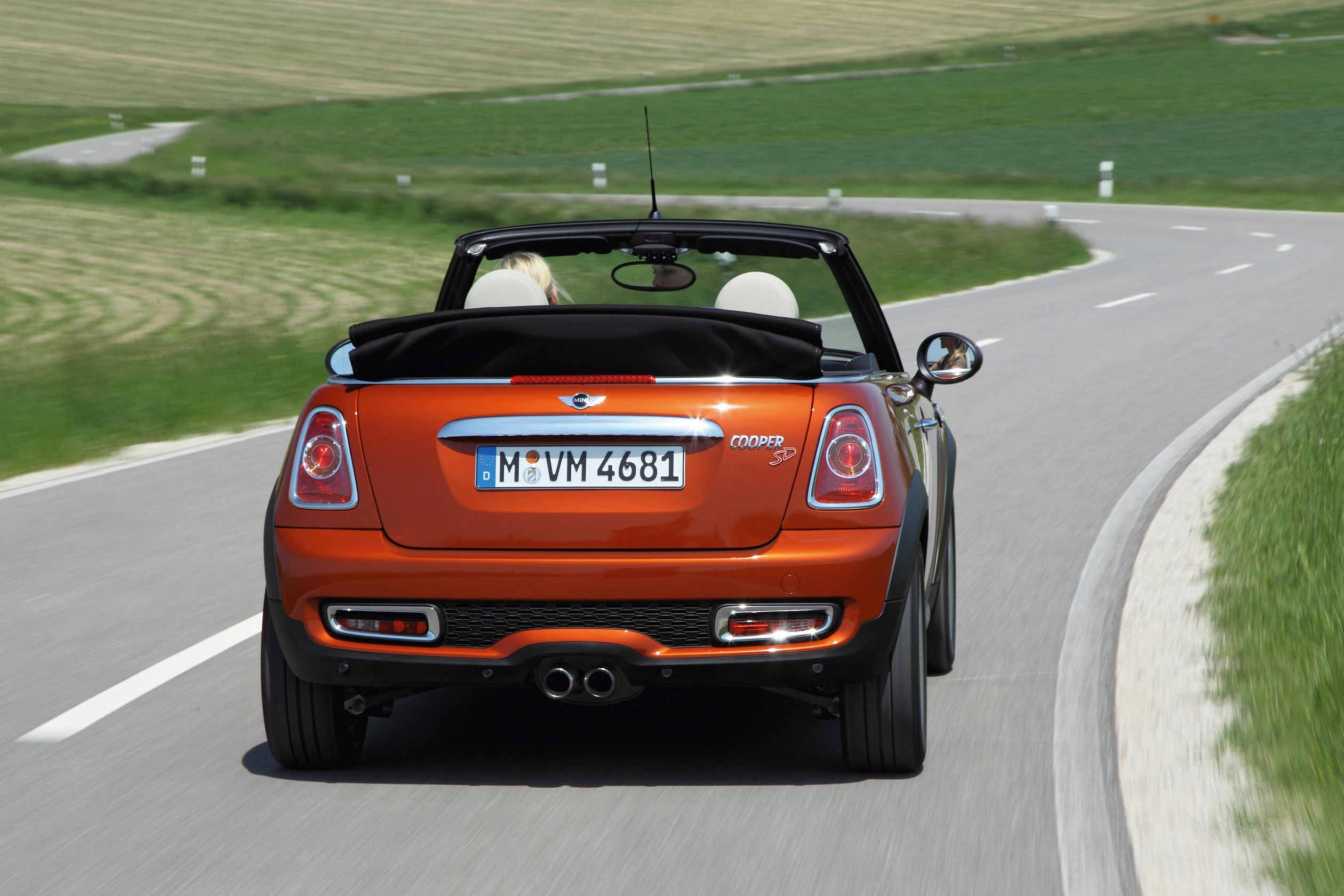 Ein orangener Cabrio-Mini Cooper fährt auf einer kurvigen Landstraße bei sonnigem Wetter. Im Hintergrund Felder und grünes Gras.
