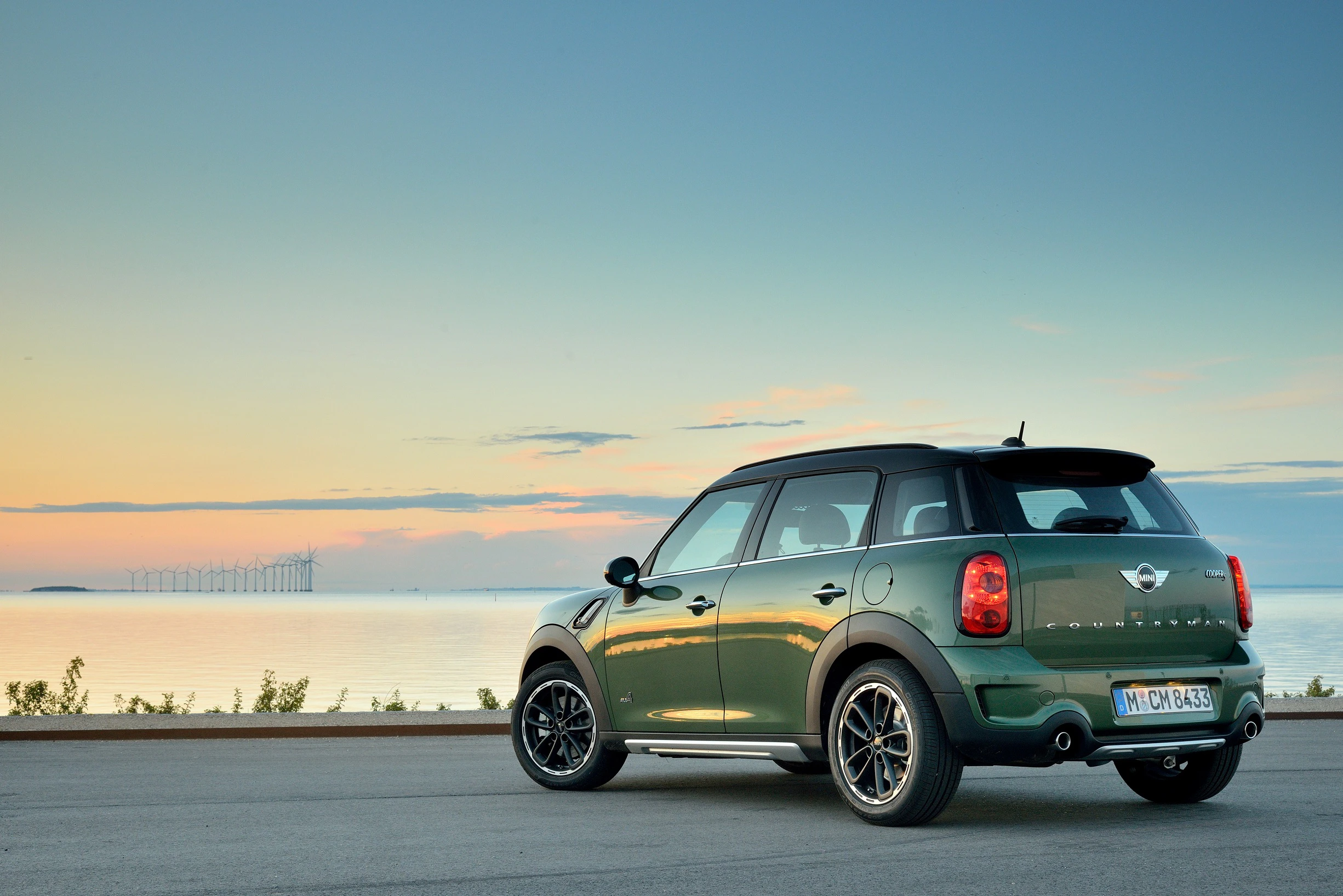 Grüner Mini Cooper steht auf Straße vor einem ruhigen See bei Sonnenuntergang, mit einer Brücke im Hintergrund.