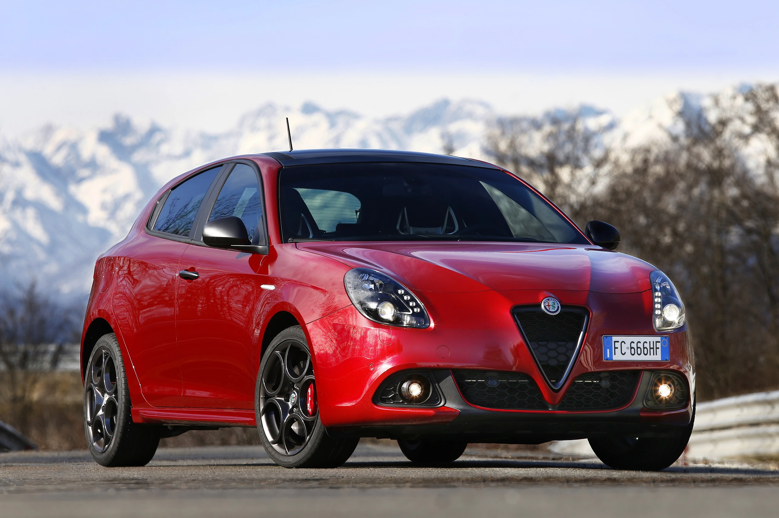 Roter Alfa Romeo Giulietta vor schneebedeckten Bergen auf einer Straße bei klarem Himmel.