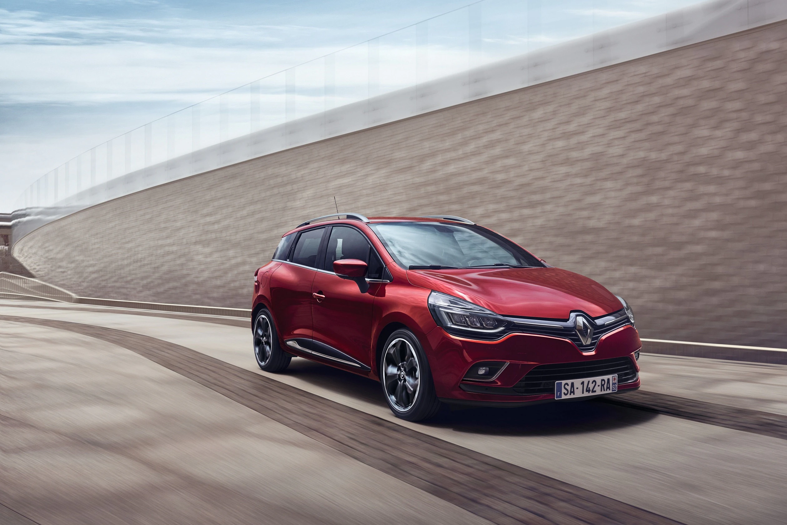 Roter Renault Clio fährt auf einer Straße mit modernem, geschwungenem Architekturrücken, unter bewölktem Himmel.