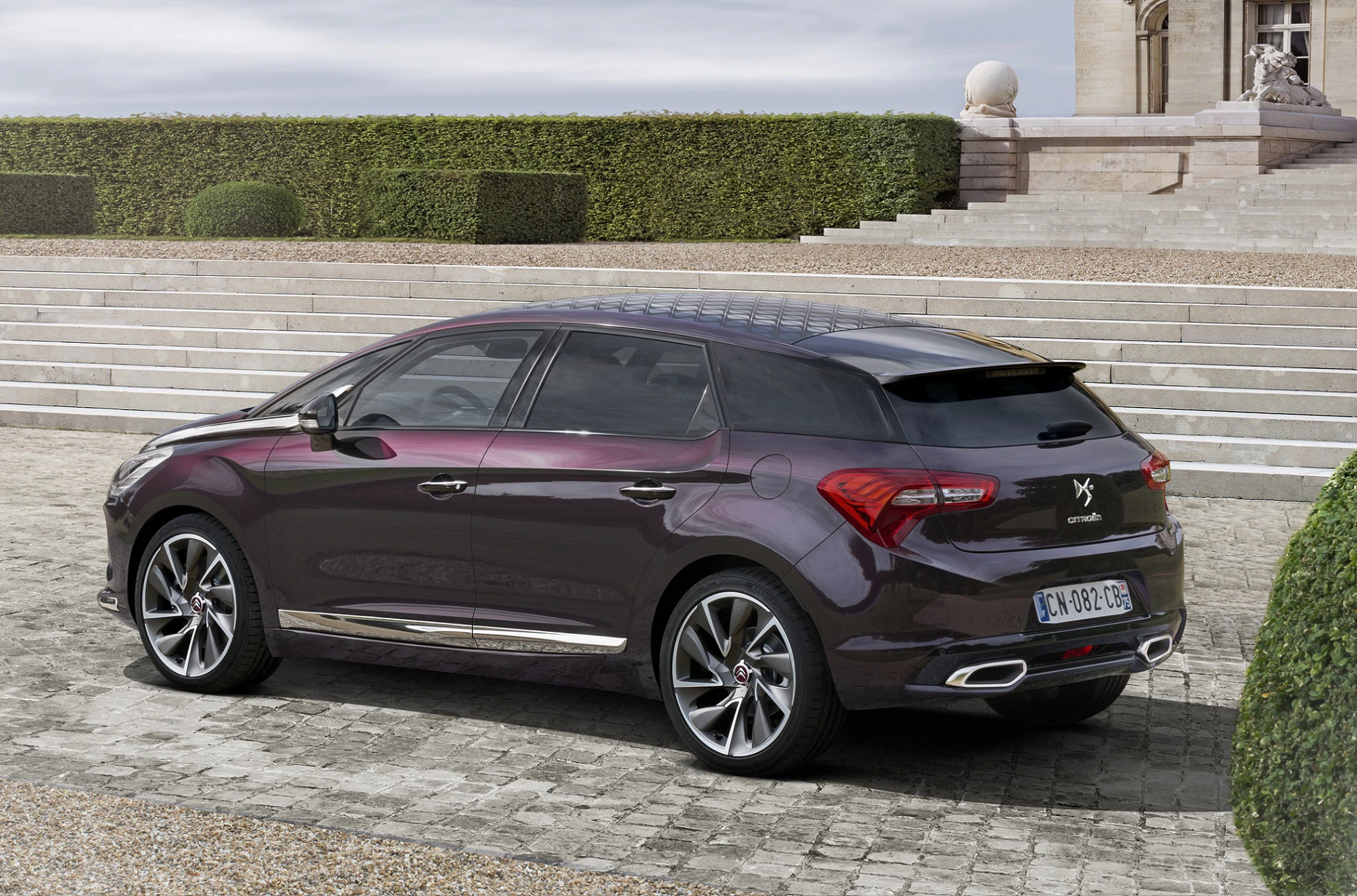 Lila Citroën DS5 parkt auf gepflastertem Platz vor einer Treppe, mit Bäumen und Hecken im Hintergrund.