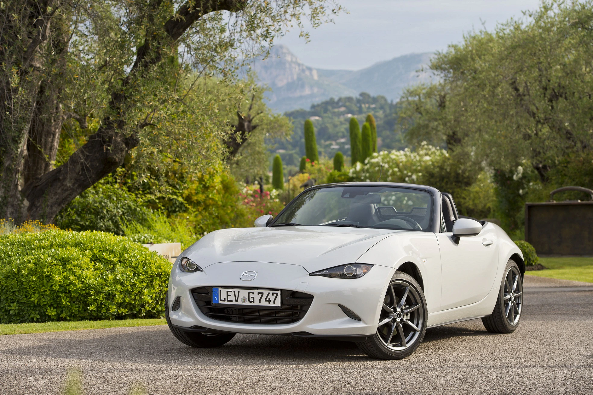 Ein weißer Mazda MX-5 Cabrio steht auf einer Straße vor einer grünen Landschaft mit Hügeln im Hintergrund.