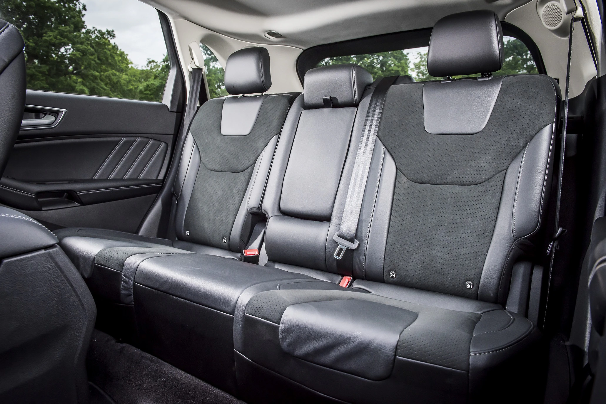 Rückbank eines Autos mit drei schwarzen Ledersitzen und Sicherheitsgurten, umgeben von großen Fenstern mit Baumansicht im Hintergrund.