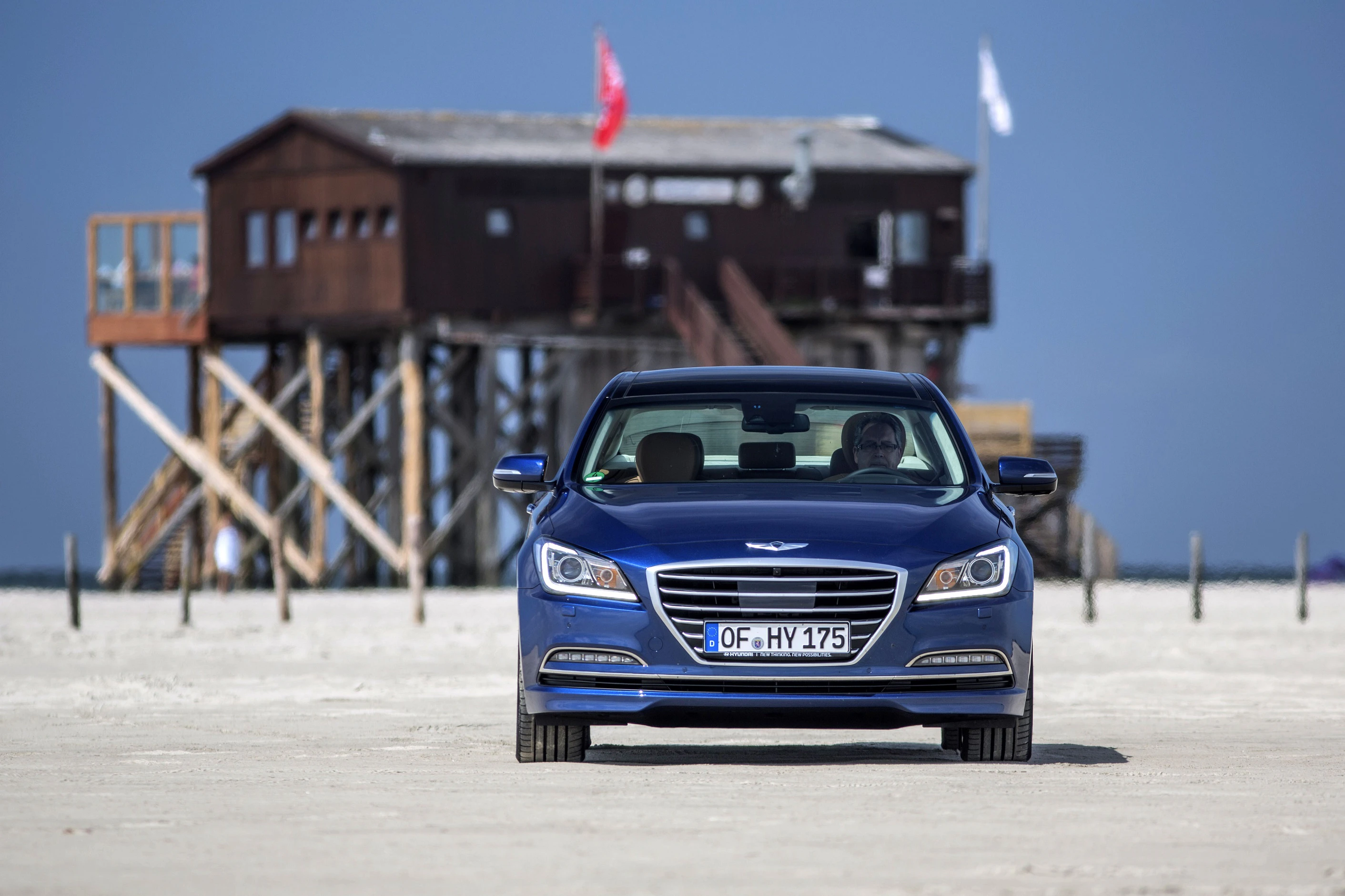 Blaues Auto auf Sandstrand vor Holzhütte mit Pfählen, blauer Himmel im Hintergrund.