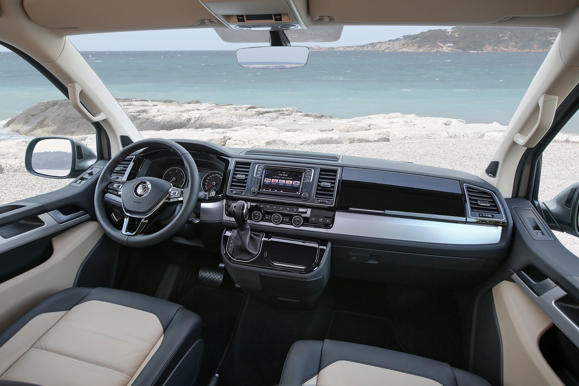 Innenraum eines Autos mit Blick aufs Meer. Moderne Ausstattung mit Lenkrad, Display und Sitzen in Beige und Schwarz.