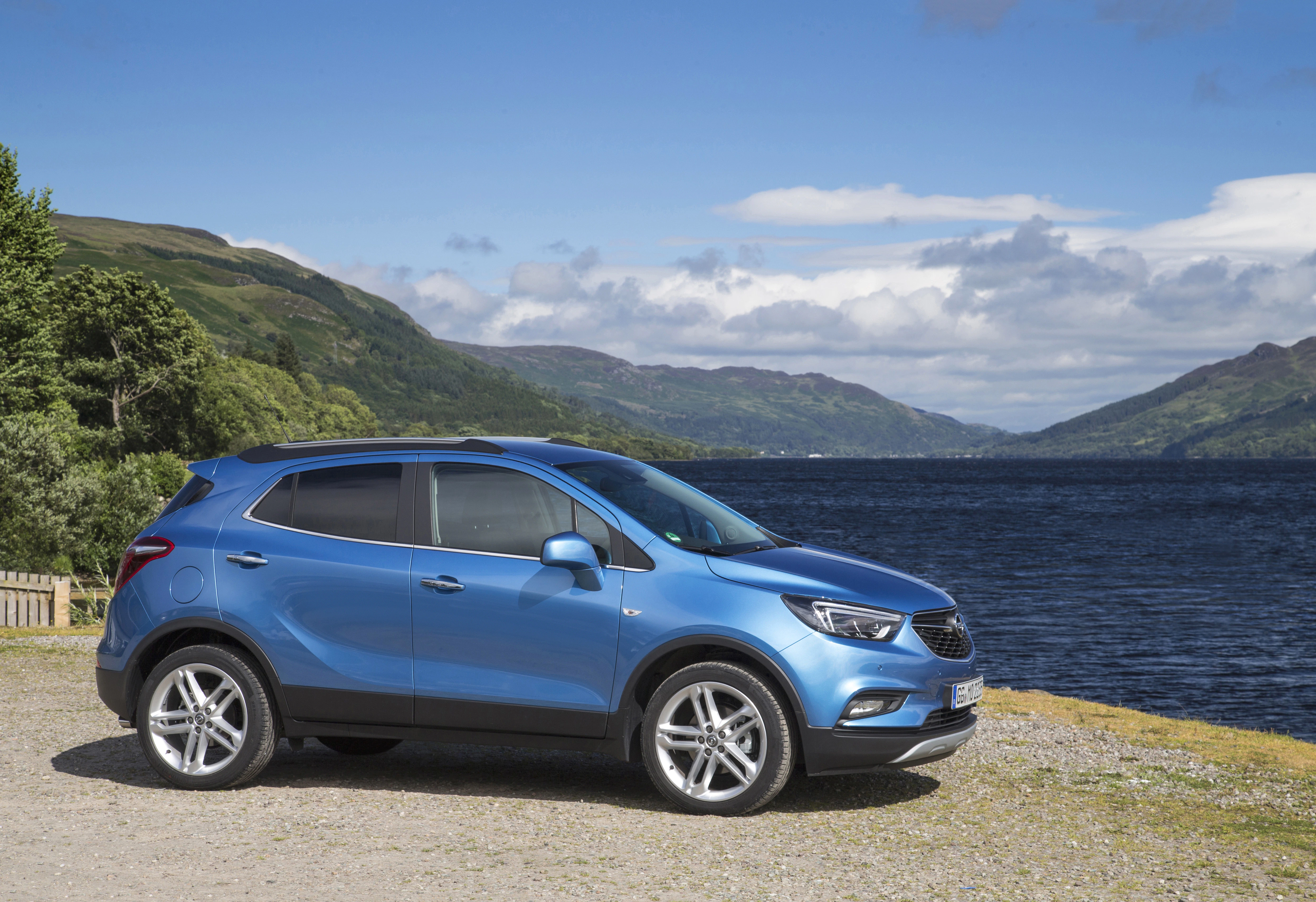 Blauer SUV parkt vor einer malerischen Kulisse mit See und grünen Hügeln unter einem klaren blauen Himmel.