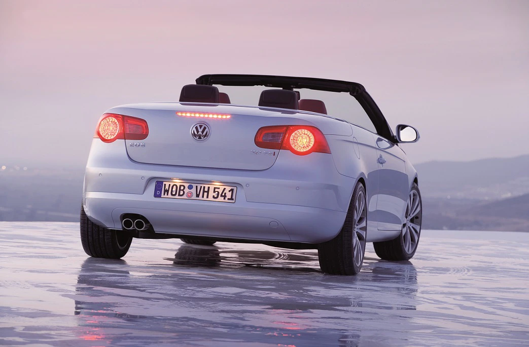 Weißes Volkswagen Cabrio mit geöffnetem Verdeck, betrachtet von hinten, auf nasser, reflektierender Fläche bei Sonnenuntergang.