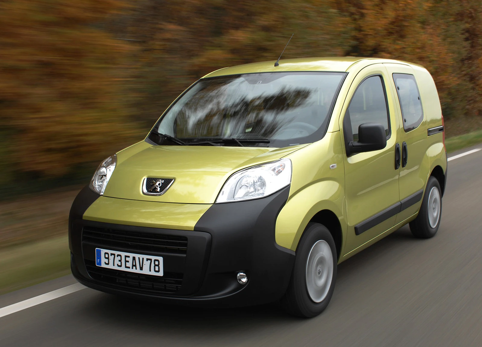Gelber Peugeot-Kastenwagen fährt auf einer Landstraße, im Hintergrund herbstliche Bäume.