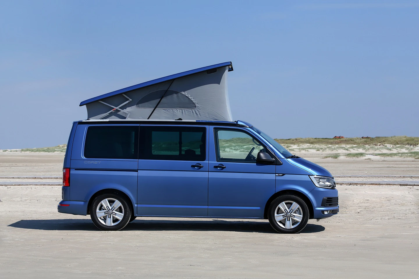 Blauer Campervan mit hochgeklapptem Dach an einem sandigen Strand vor einem klaren Himmel.
