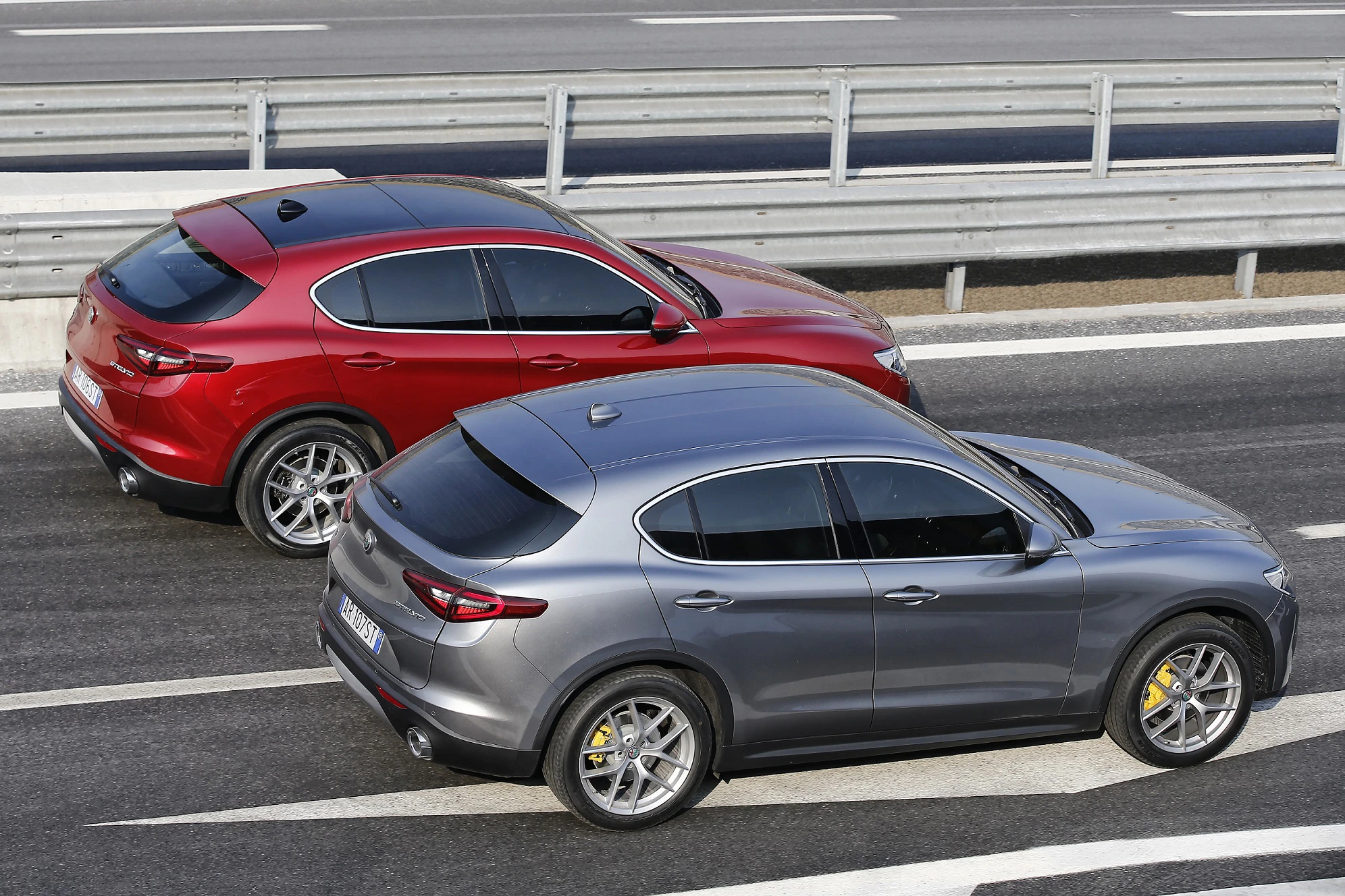 Zwei SUV fahren auf einer mehrspurigen Autobahn, einer in Rot, der andere in Grau. Sie sind von einer Barriere aus Metall umgeben.