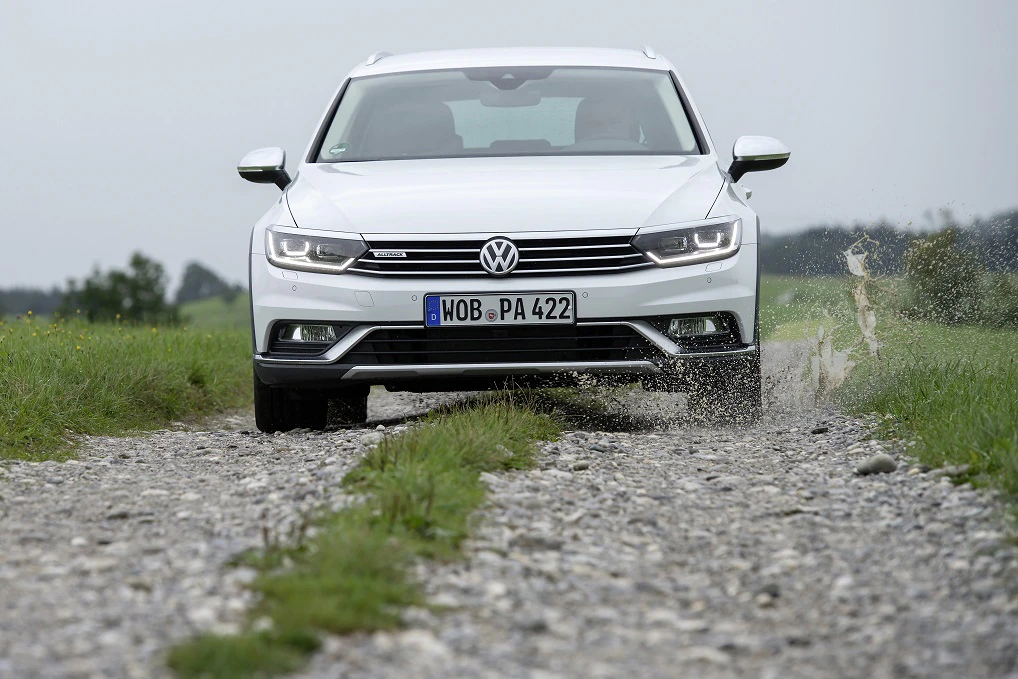Weißer Volkswagen Passat fährt auf einer steinigen, matschigen Straße durch eine Landschaft, Spritzwasser an den Reifen.