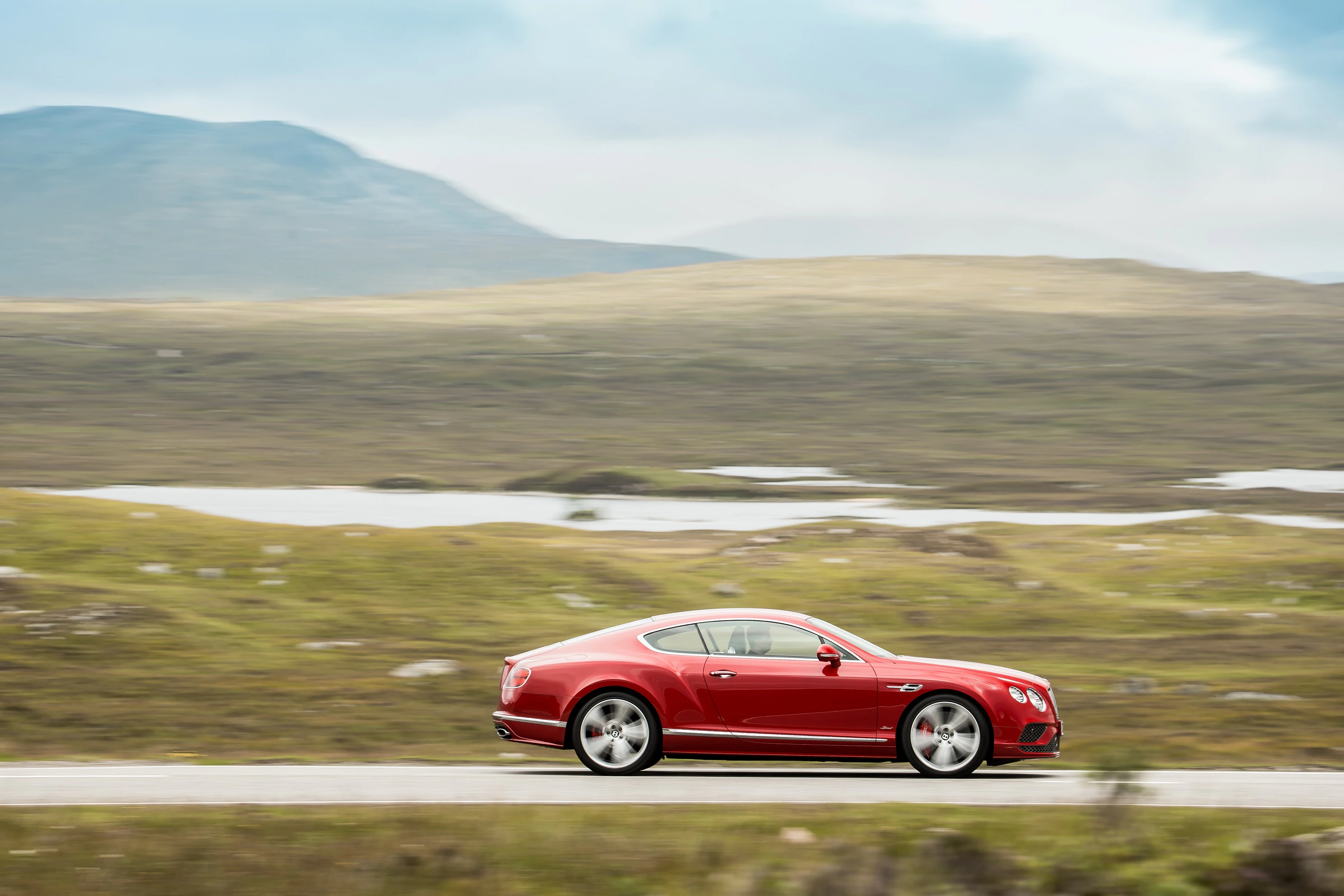 Rotes Auto fährt schnell auf einer Straße durch eine hügelige Landschaft unter bewölktem Himmel.