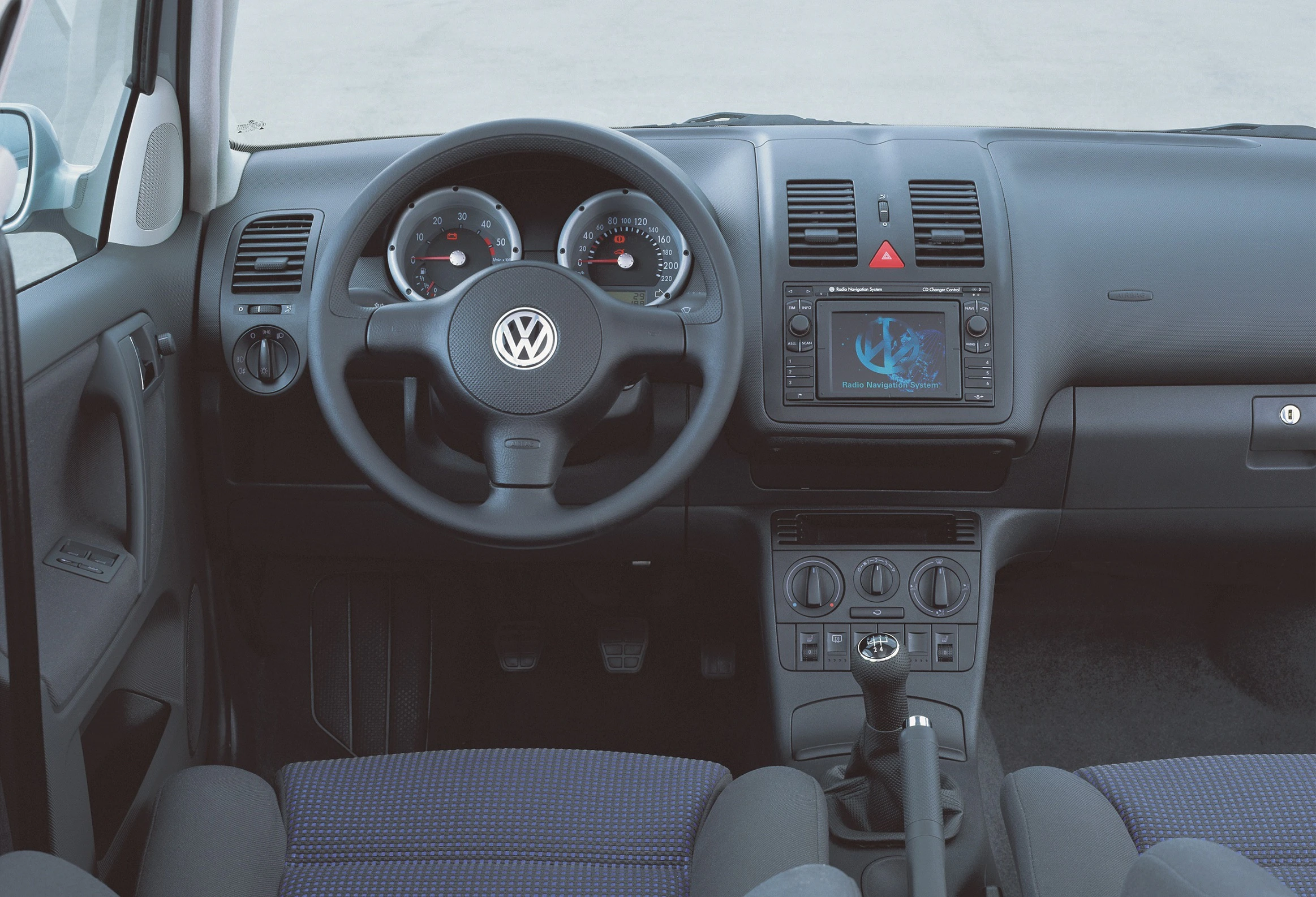 Innenraum eines Volkswagen mit Lenkrad, Armaturenbrett, Tacho und zentralem Bildschirm. Schaltknüppel und Kontrollpanel sind sichtbar.