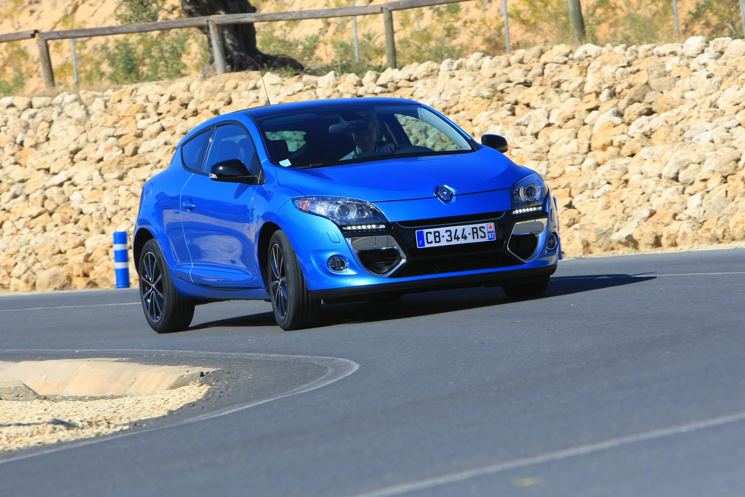 Blauer Sportwagen fährt auf einer kurvigen Straße entlang einer Steinmauer, schöner sonniger Tag, klarer Himmel im Hintergrund.