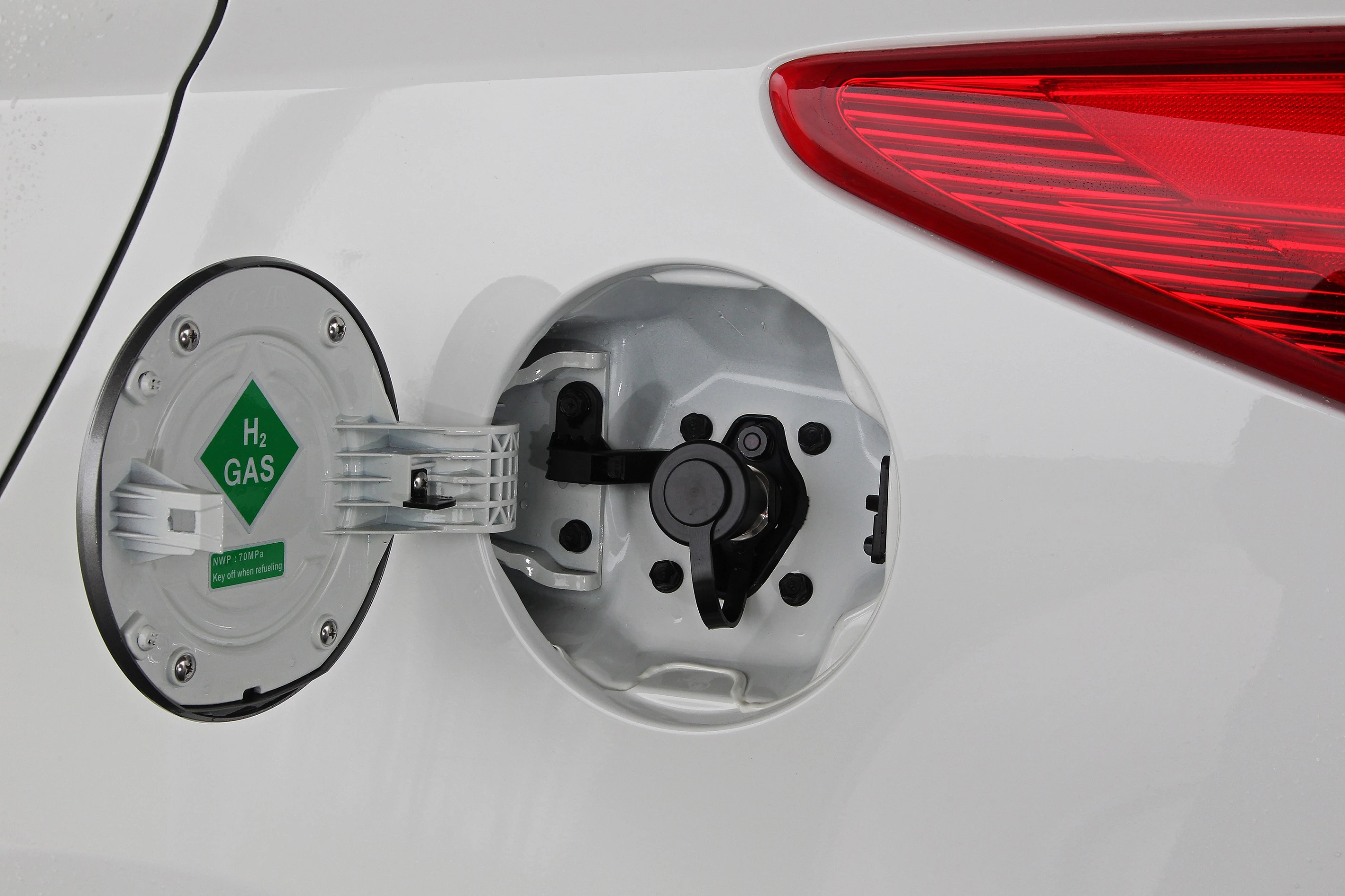 Nahaufnahme eines geöffneten Wasserstofftankdeckels an einem weißen Auto, mit einem H₂-GAS-Symbol und einem roten Rücklicht im Hintergrund.