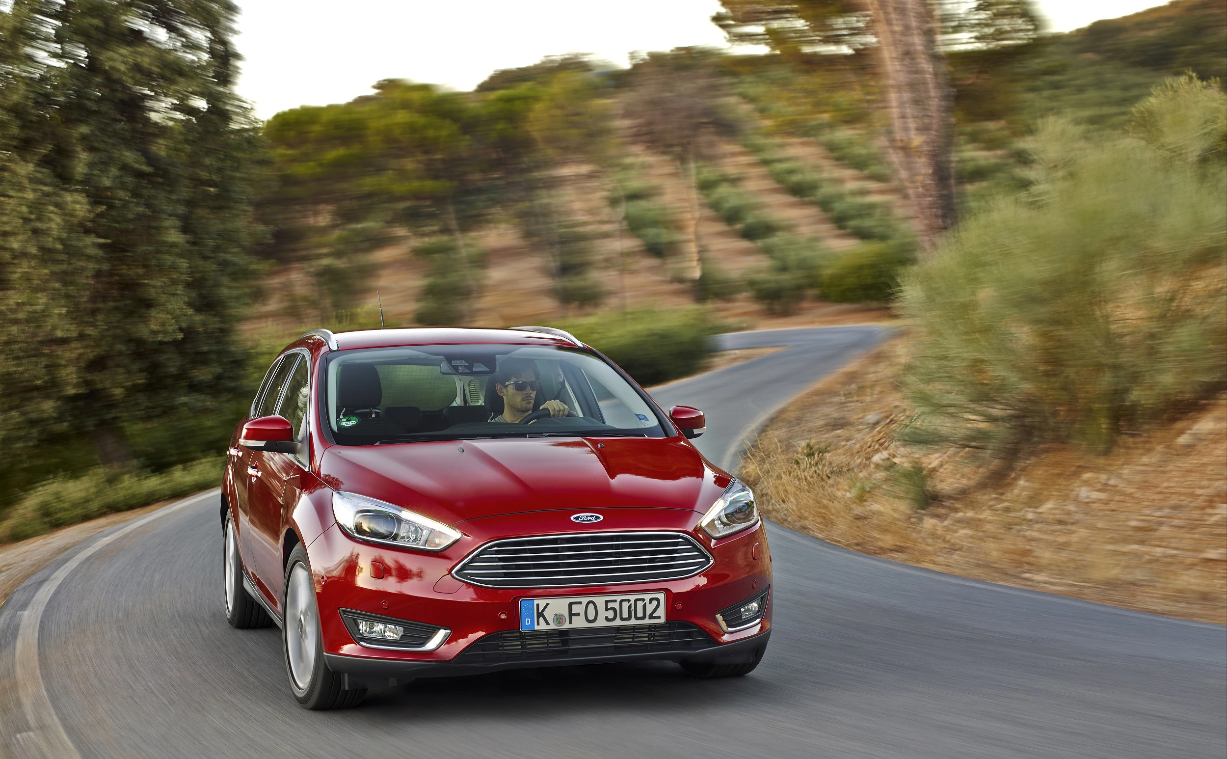 Roter Ford Focus fährt auf kurviger Landstraße, umgeben von Bäumen und Hügeln.