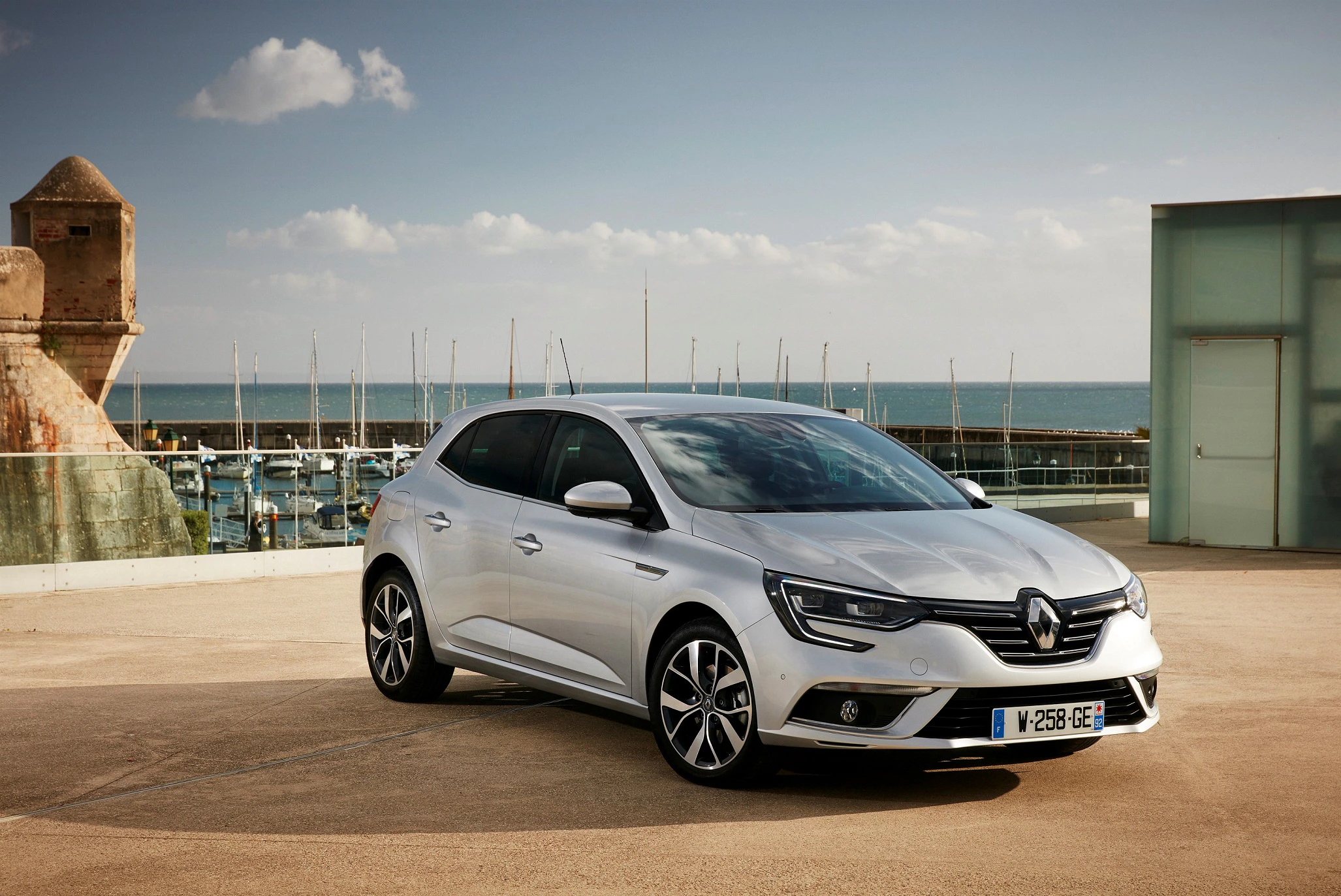 Silberner Renault auf einem Parkplatz am Meer, im Hintergrund ein Yachthafen und bewölkter Himmel.