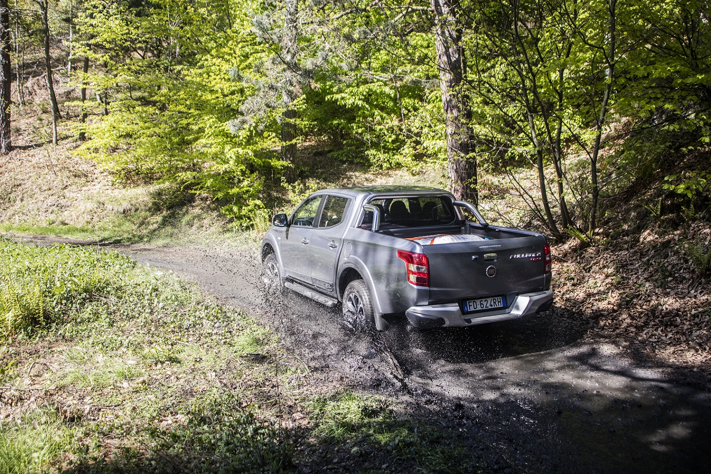 Silberner Pick-up fährt durch matschigen Waldweg, spritzt Wasser auf, umgeben von grünen Bäumen und Lichtungen im sonnigen Frühling.