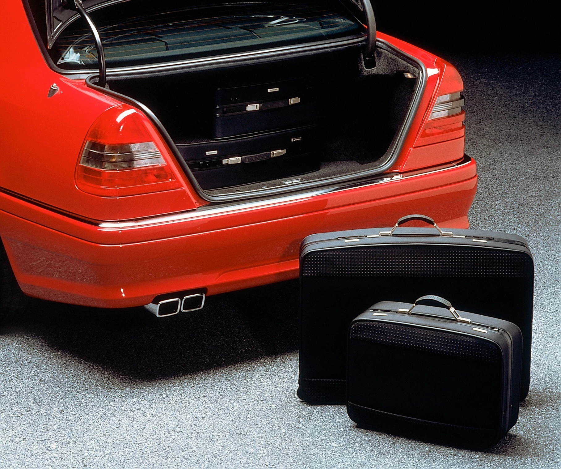 Roter Kofferraum eines Autos mit geöffnetem Heck, innen zwei Koffer. Zwei weitere schwarze Koffer stehen auf dem Boden daneben.