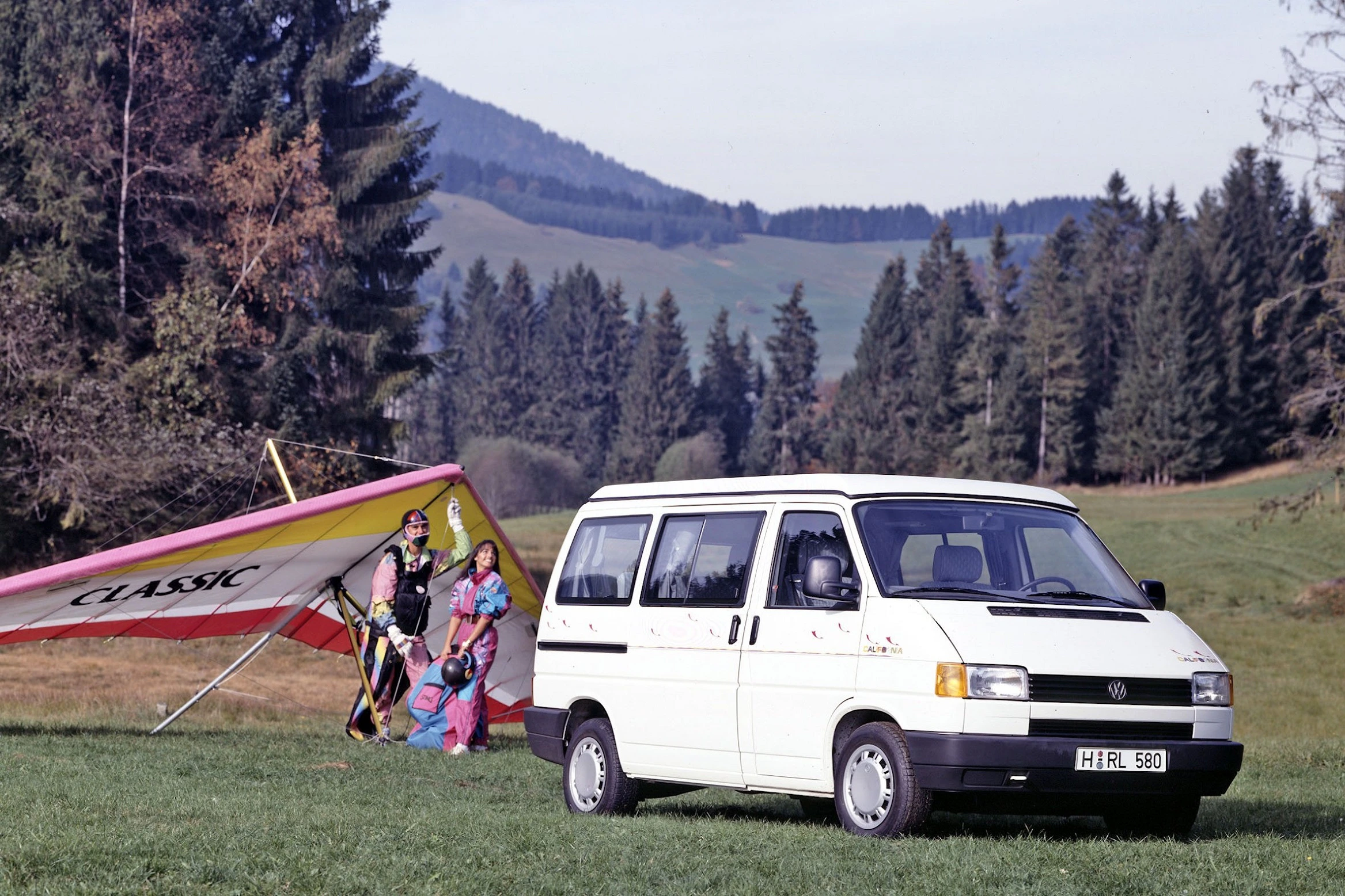 Weißer VW-Bus in ländlicher Landschaft. Im Hintergrund stehen Personen mit einem bunt gestreiften Hängegleiter.