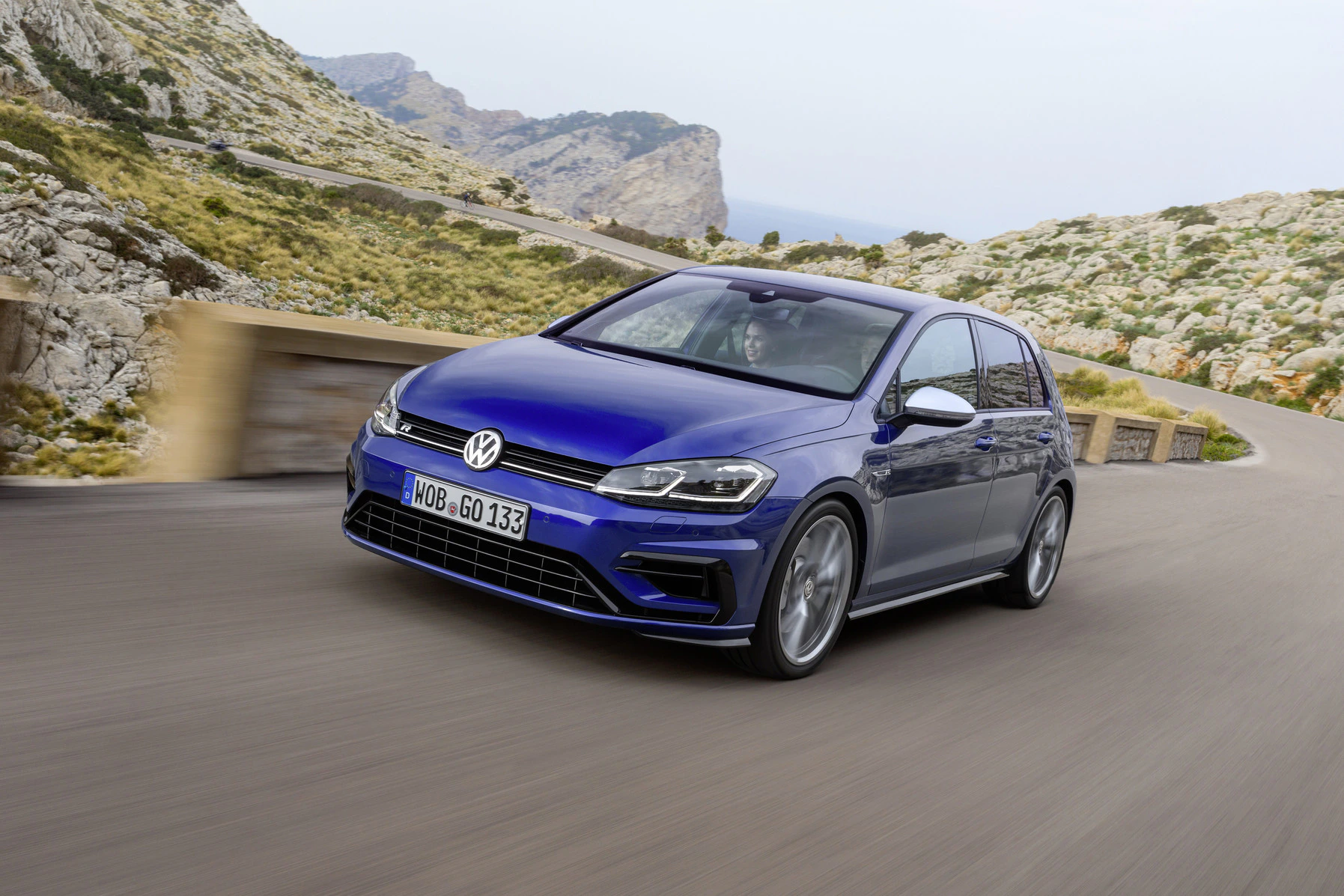Ein blauer Volkswagen Golf fährt auf einer kurvenreichen Bergstraße. Im Hintergrund sind Felsen und Vegetation sichtbar.
