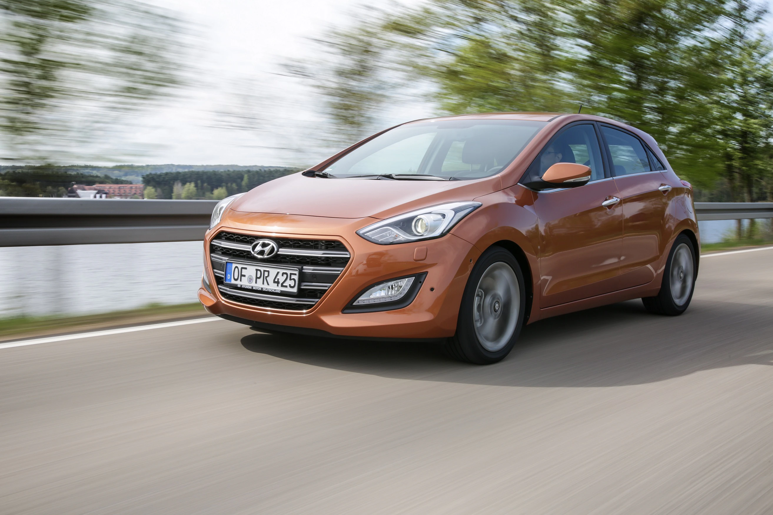 Ein orangefarbener Hyundai i30 fährt dynamisch auf einer Landstraße, Bäume im Hintergrund verschwommen, Autofahren bei Tageslicht.