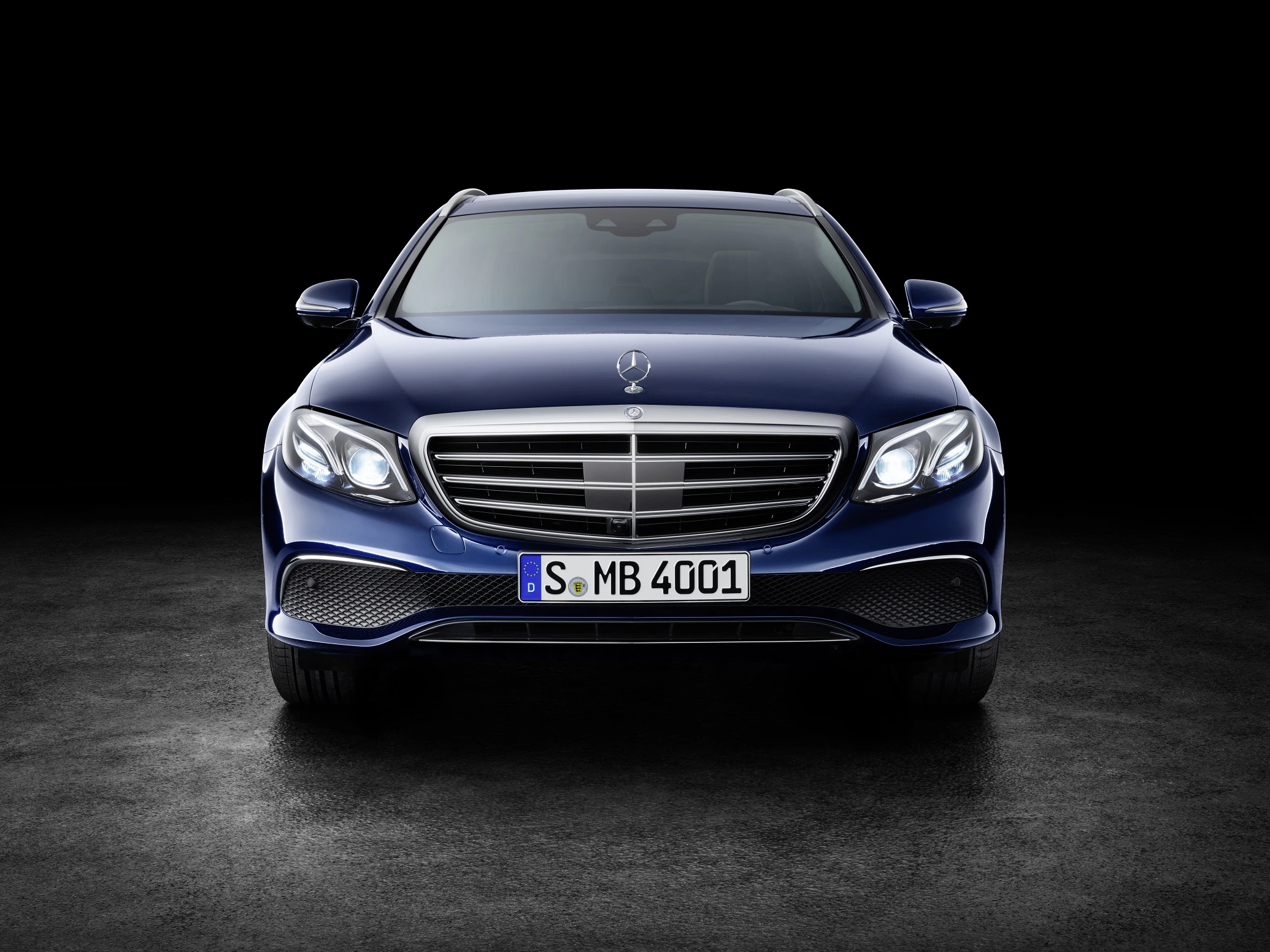 Blauer Mercedes-Benz E-Klasse Kombi, Frontansicht auf dunklem Hintergrund. Elegantes Design mit markantem Kühlergrill und LED-Scheinwerfern.