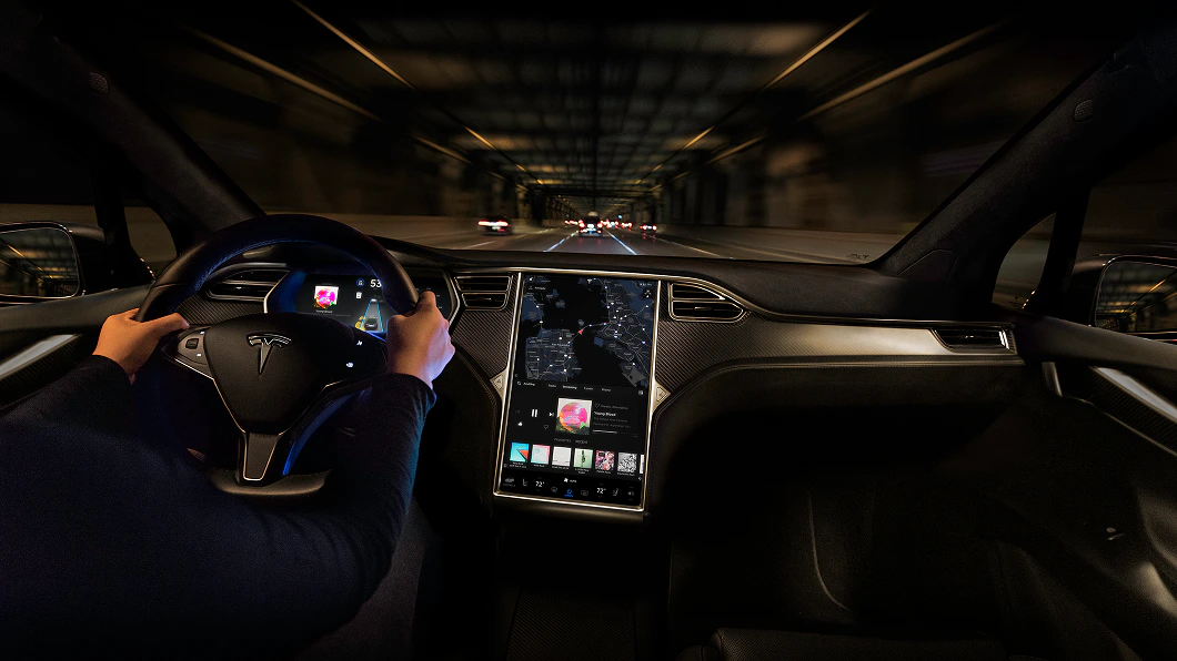 Innenraum eines Tesla-Autos bei Nachtfahrt. Fahrer hält das Lenkrad, großes zentrales Display zeigt Karten- und Unterhaltungsapps.
