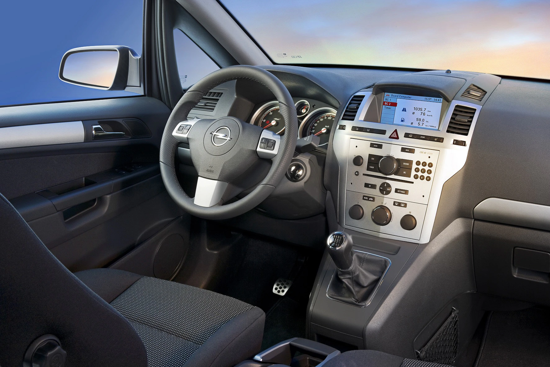Innenraum eines Autos mit Lenkrad, Schalthebel und Instrumententafel. Moderne Bedienelemente und graue Polsterung.