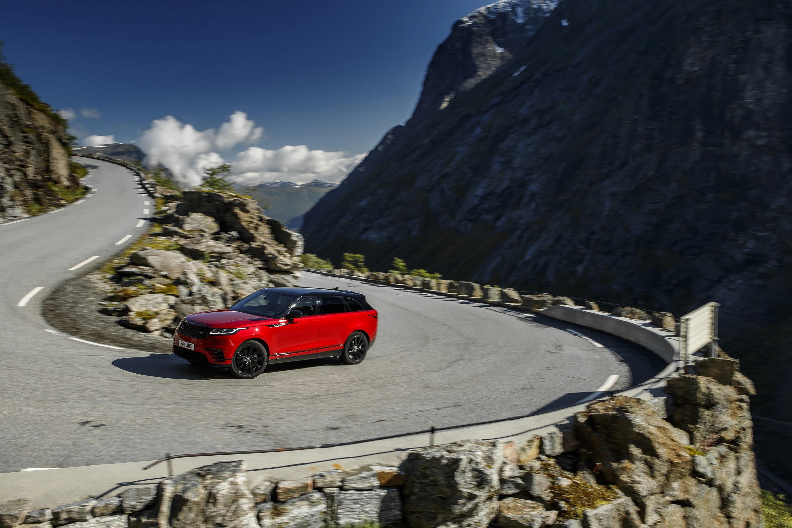 Rotes Auto fährt auf kurviger Bergstraße entlang felsiger Landschaft mit Bergen und blauem Himmel im Hintergrund.