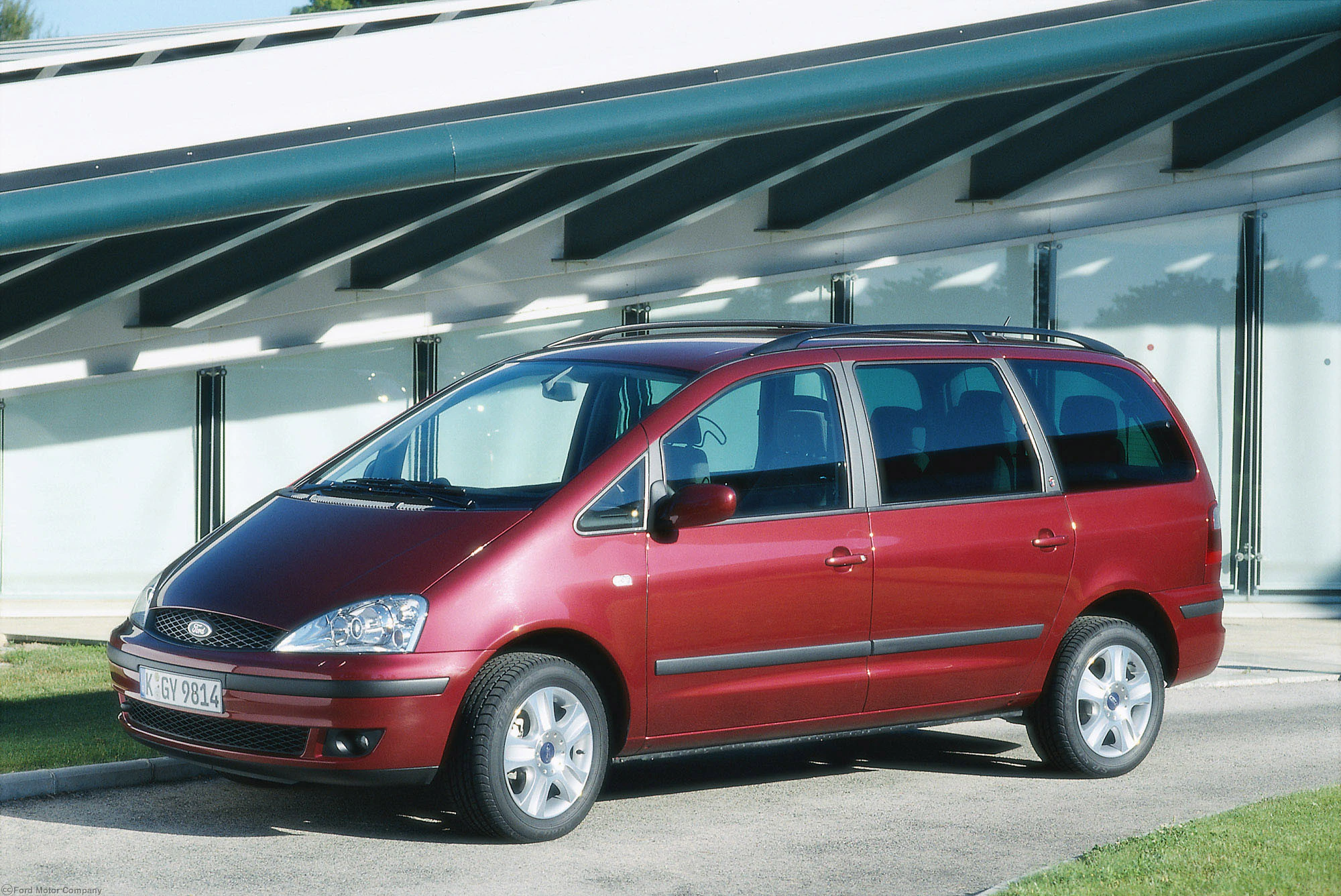 Roter Ford Galaxy Minivan parkt vor einem modernen weißen Gebäude mit Glasfenstern.