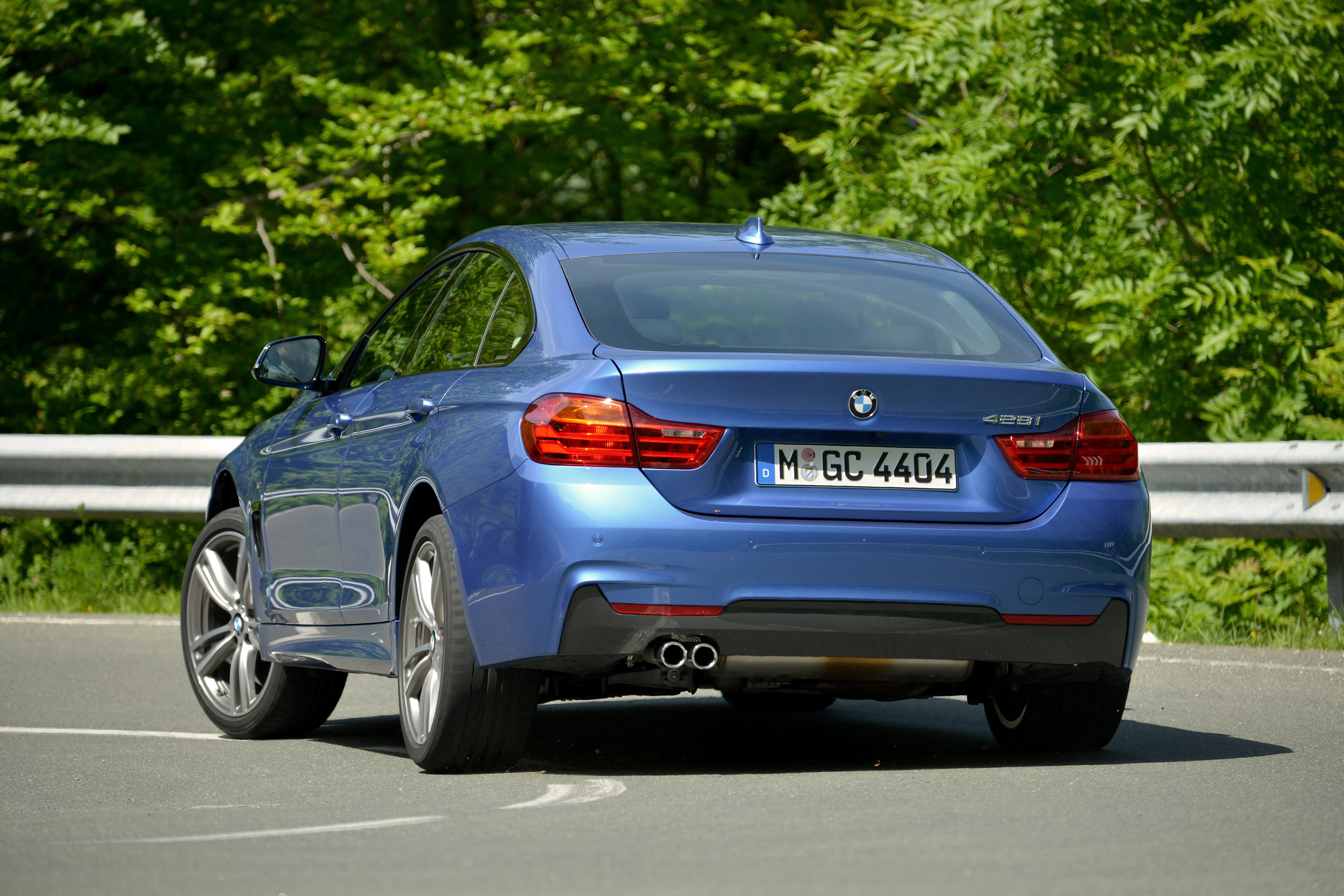 Blauer BMW 4er Gran Coupé auf einer Straße, umgeben von grünen Bäumen. Heckansicht mit klarem Nummernschild und Doppelauspuff.