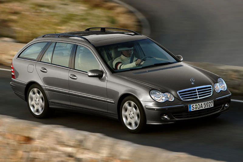 Silberner Mercedes-Benz Kombi fährt eine kurvige Straße entlang. Fahrer trägt einen Hut. Auto ist von einer Steinmauer umgeben.