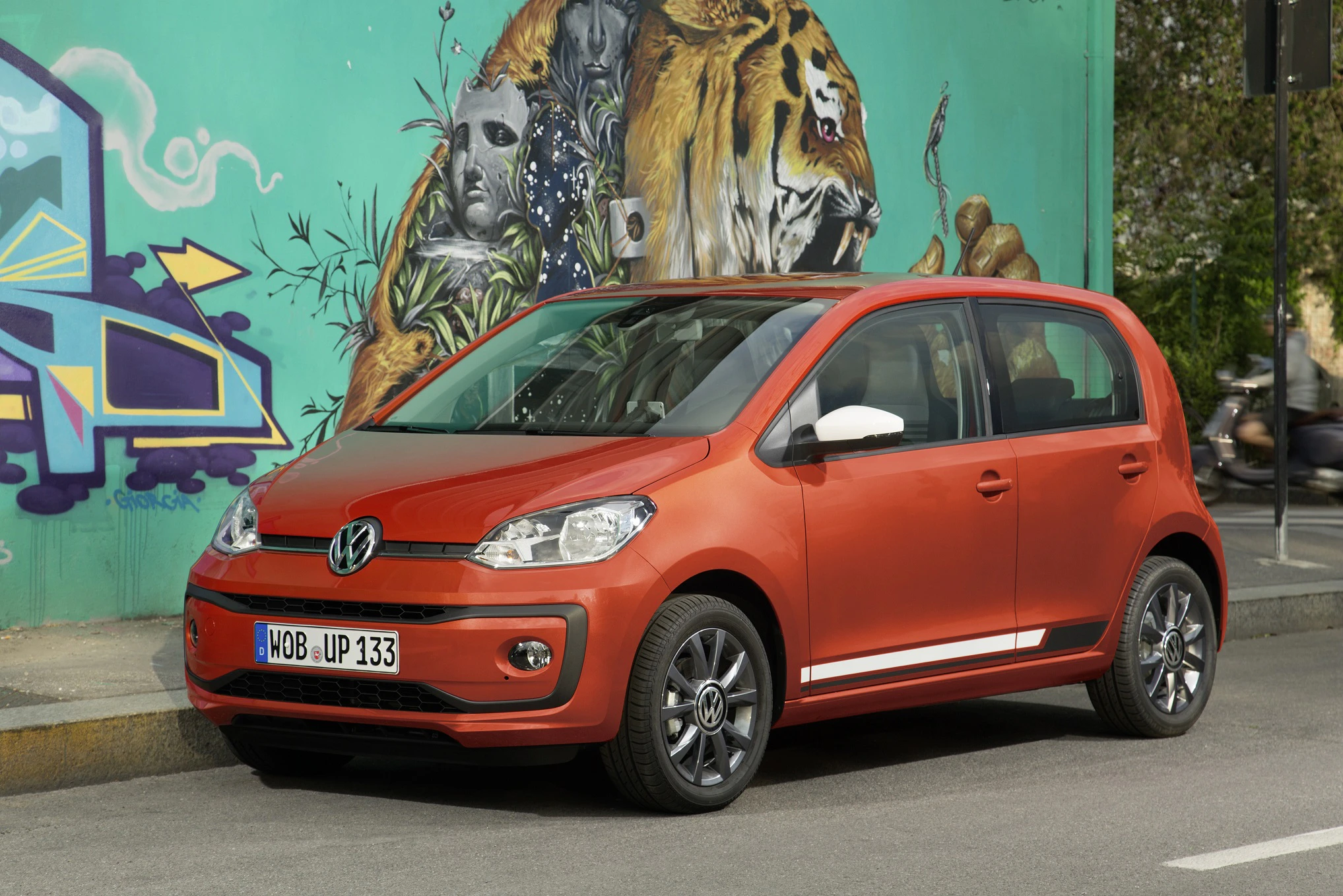 Ein roter Volkswagen Kleinwagen parkt vor einer bunten Wand mit Graffiti-Kunstwerk, das einen Löwen und eine maskierte Figur zeigt.