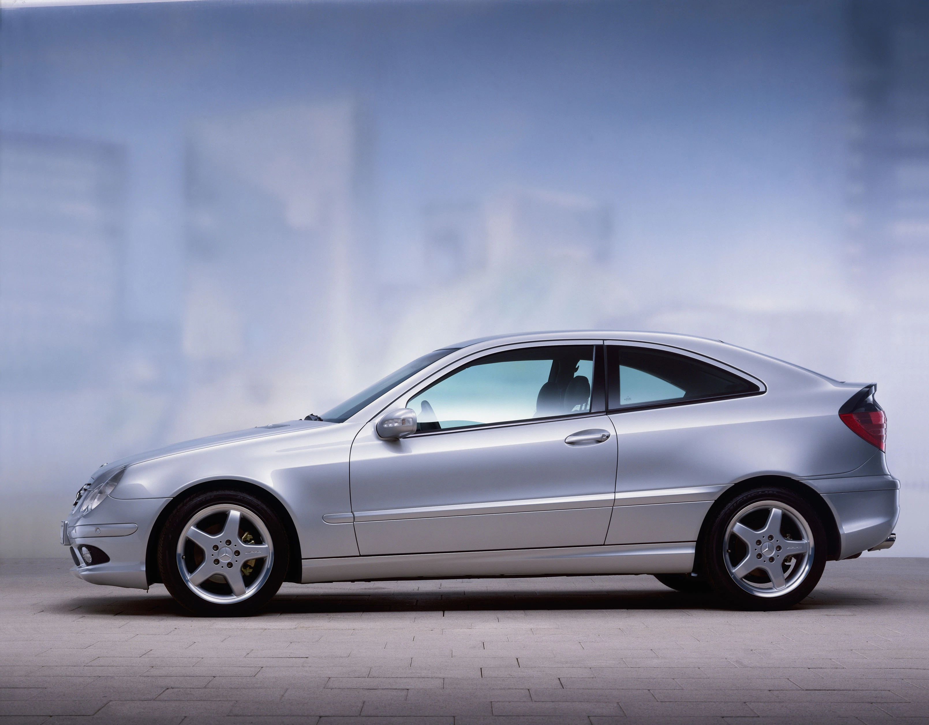 Silbernes Coupé-Auto, seitlich aufgenommen, vor einem verschwommenen Stadtbild. Elegantes Design und moderne Aluminiumfelgen.
