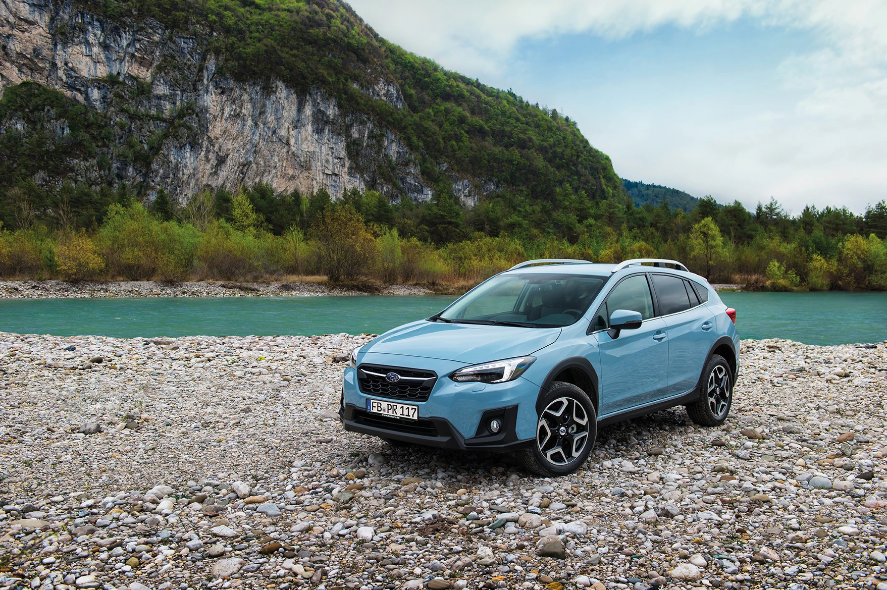 Ein blauer SUV steht auf einem steinigen Flussufer vor einer grünen, bewaldeten Berglandschaft unter bewölktem Himmel.