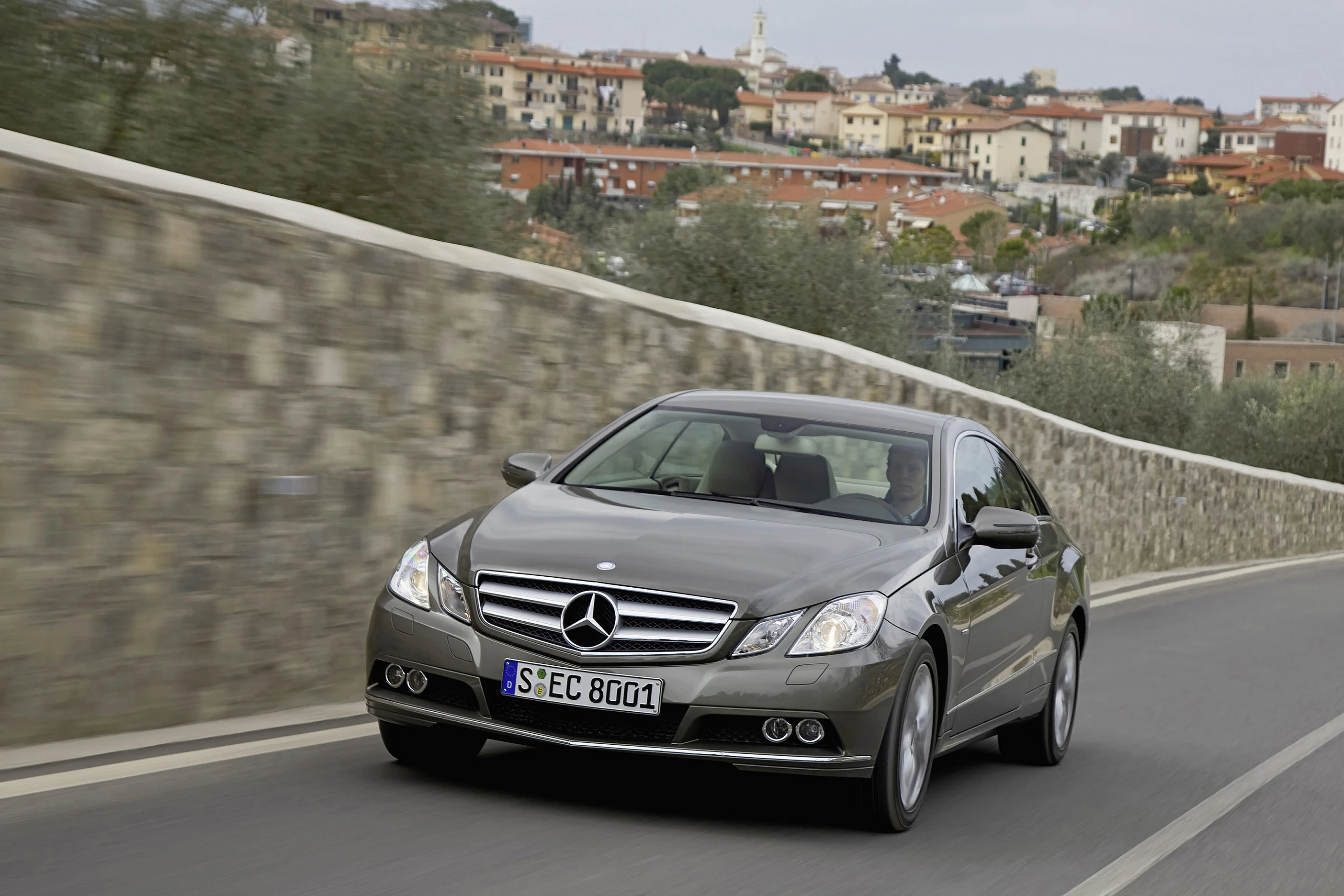 Silberner Mercedes-Benz fährt auf einer Straße neben einer Steinmauer, im Hintergrund eine mediterrane Stadtlandschaft.