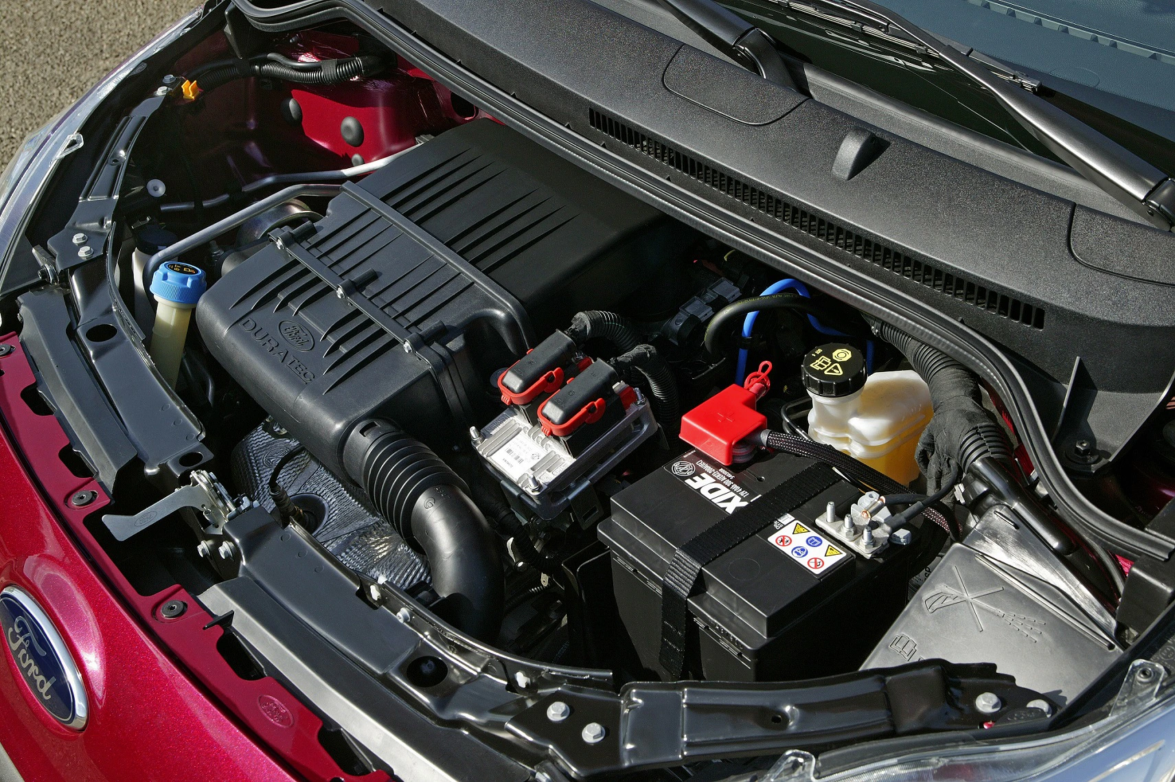 Motorraum eines Autos mit sichtbarer Batterie, Luftfilter und verschiedenen Schläuchen und Kabeln. Automarke nicht erkennbar.