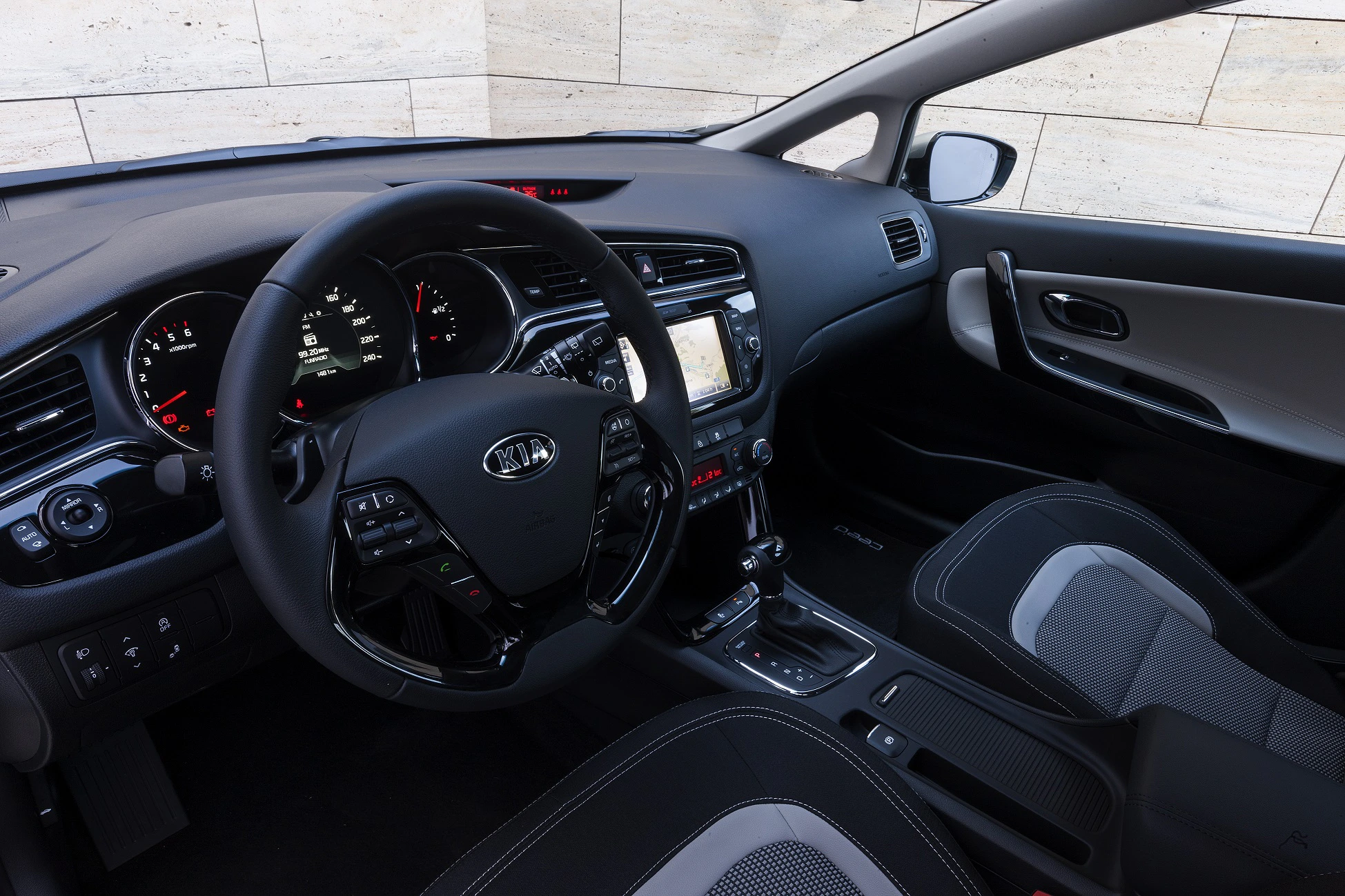 Innenraum eines modernen Autos mit Lenkrad, Tachometer und Touchscreen-Display. Schwarze Sitze mit hellgrauen Akzenten.