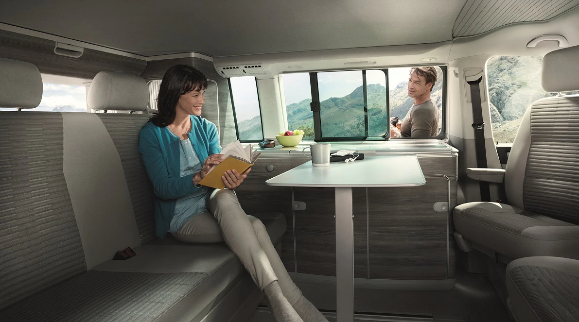 Eine Frau liest ein Buch auf einer Bank in einem Wohnmobil, während ein Mann am Fenster steht. Draußen sind Berge zu sehen.