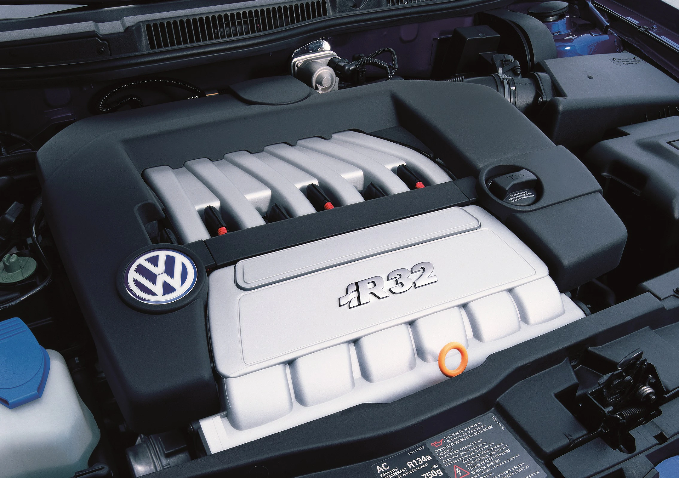Nahaufnahme eines Volkswagen-Motors mit R32-Emblem. Der Motorraum zeigt Abdeckungen und das VW-Logo.