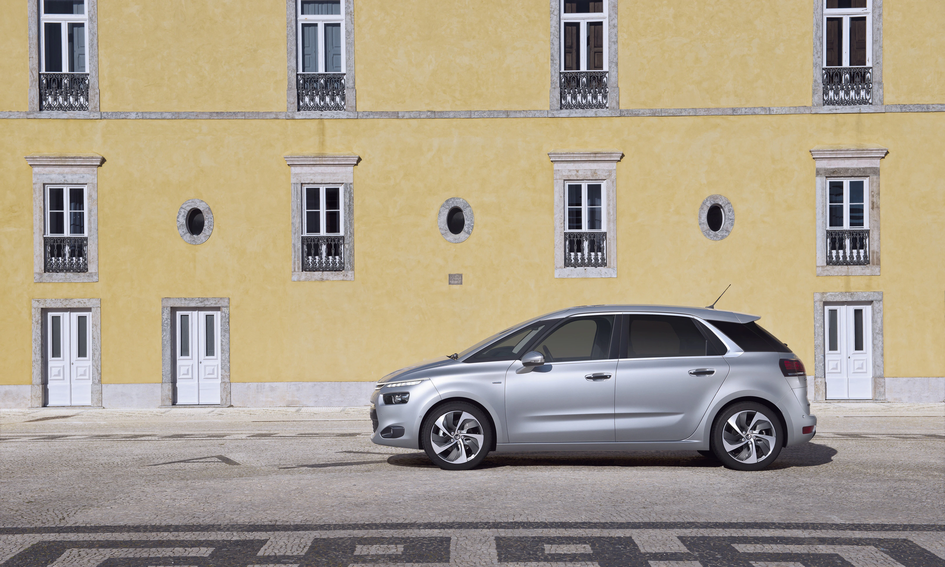 Silberner Minivan parkt vor einer gelben Gebäude mit weißen Fenstern und Türen, auf gepflastertem Boden mit Mosaikmuster.